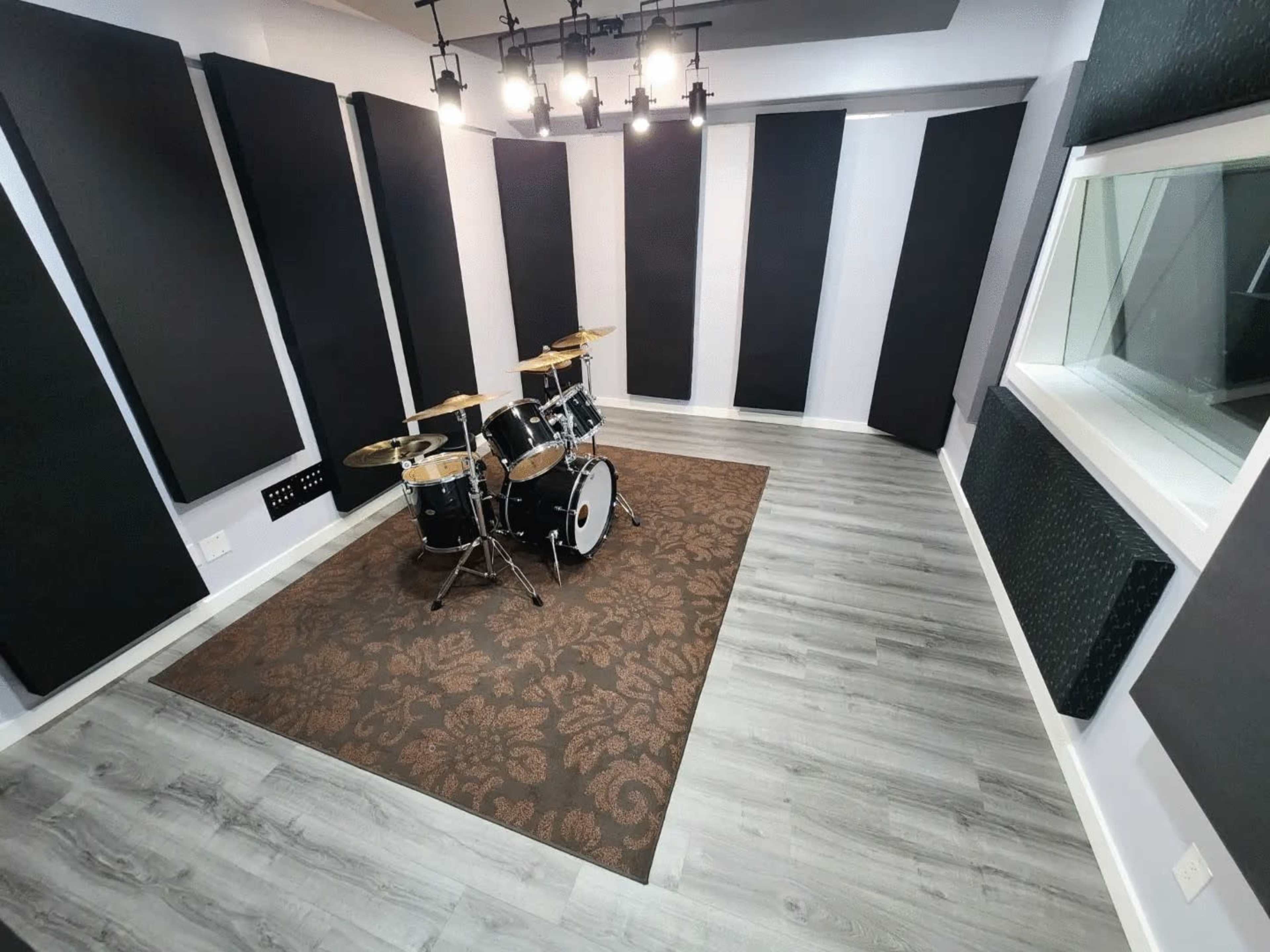 The image shows a well-equipped recording studio featuring a drum set in the center, surrounded by acoustic panels on the walls and a large window.