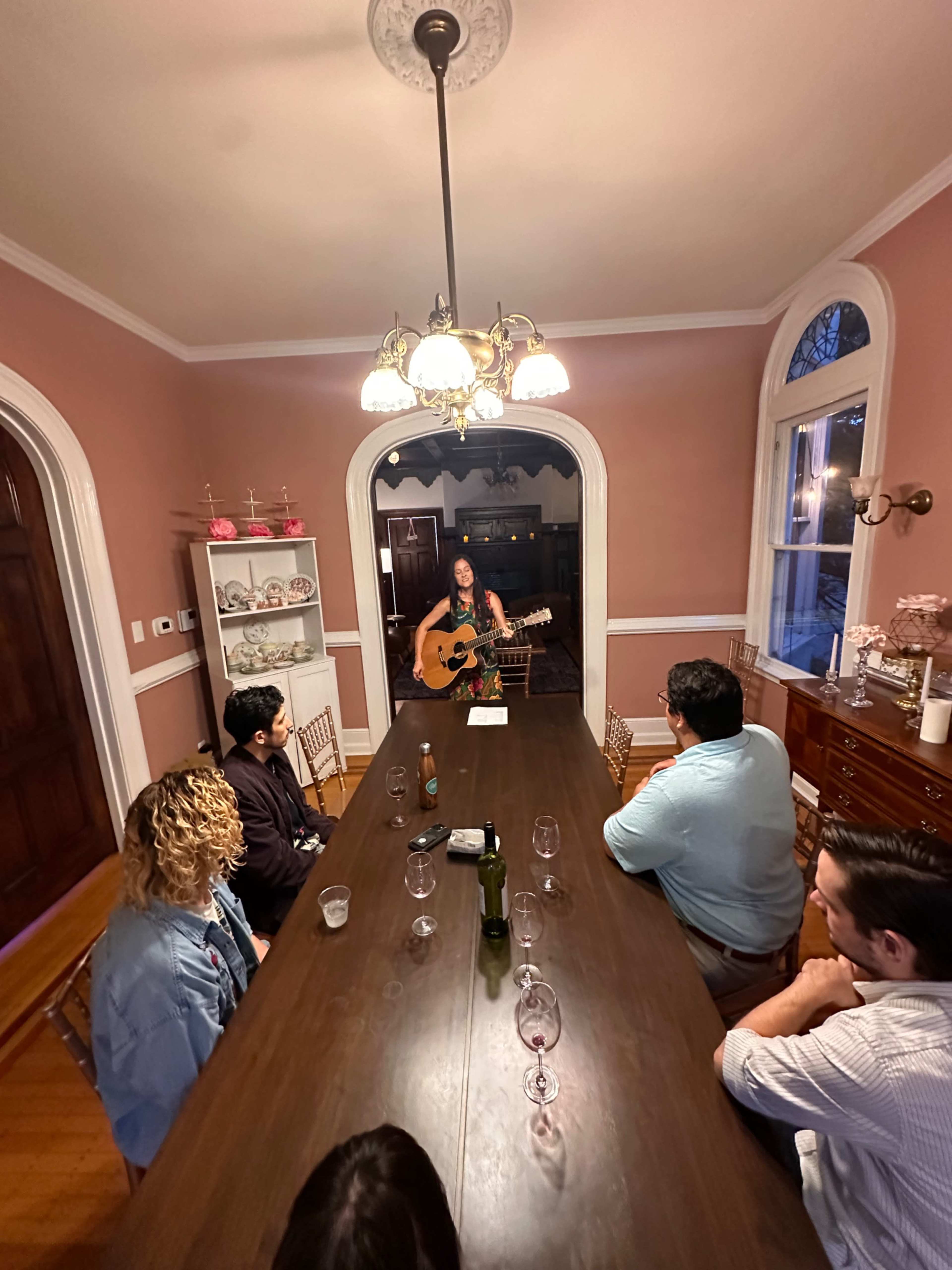 A musician plays guitar at a long dining table where several people are seated, listening attentively in a warmly lit room.