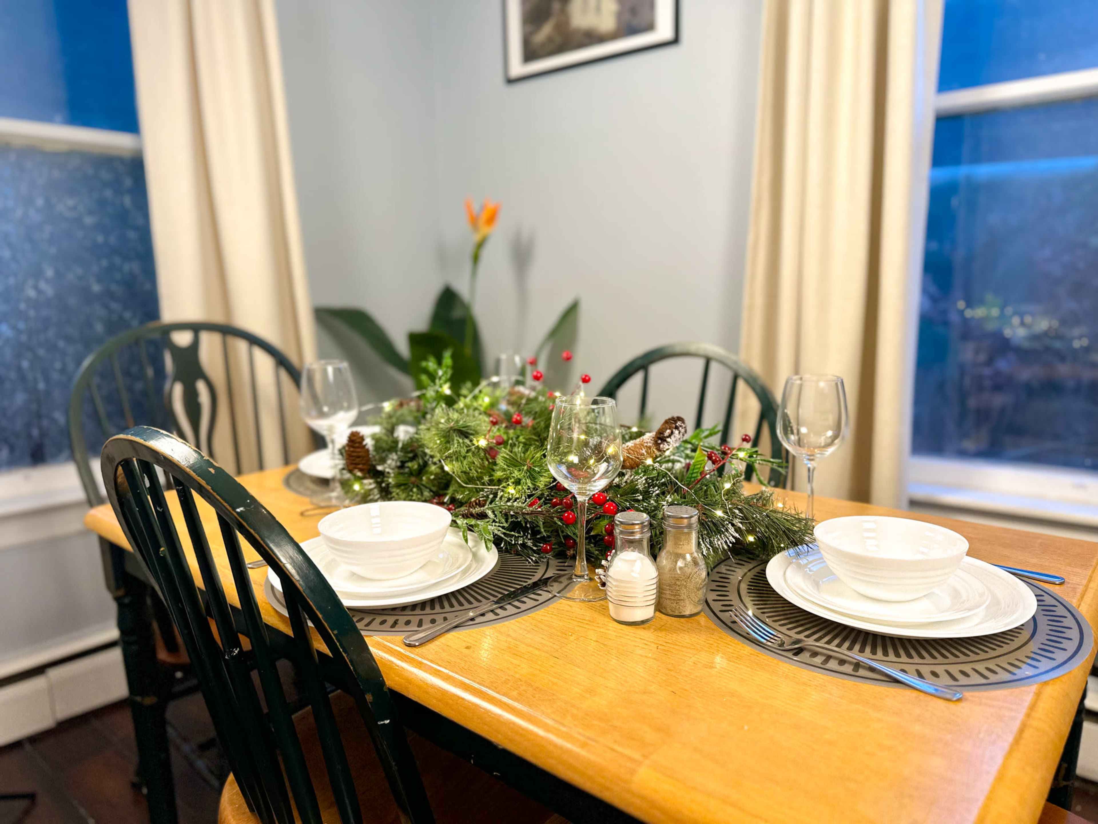 A neatly arranged dining table is set with white dishware, wine glasses, and a decorative centerpiece of greenery and pinecones.