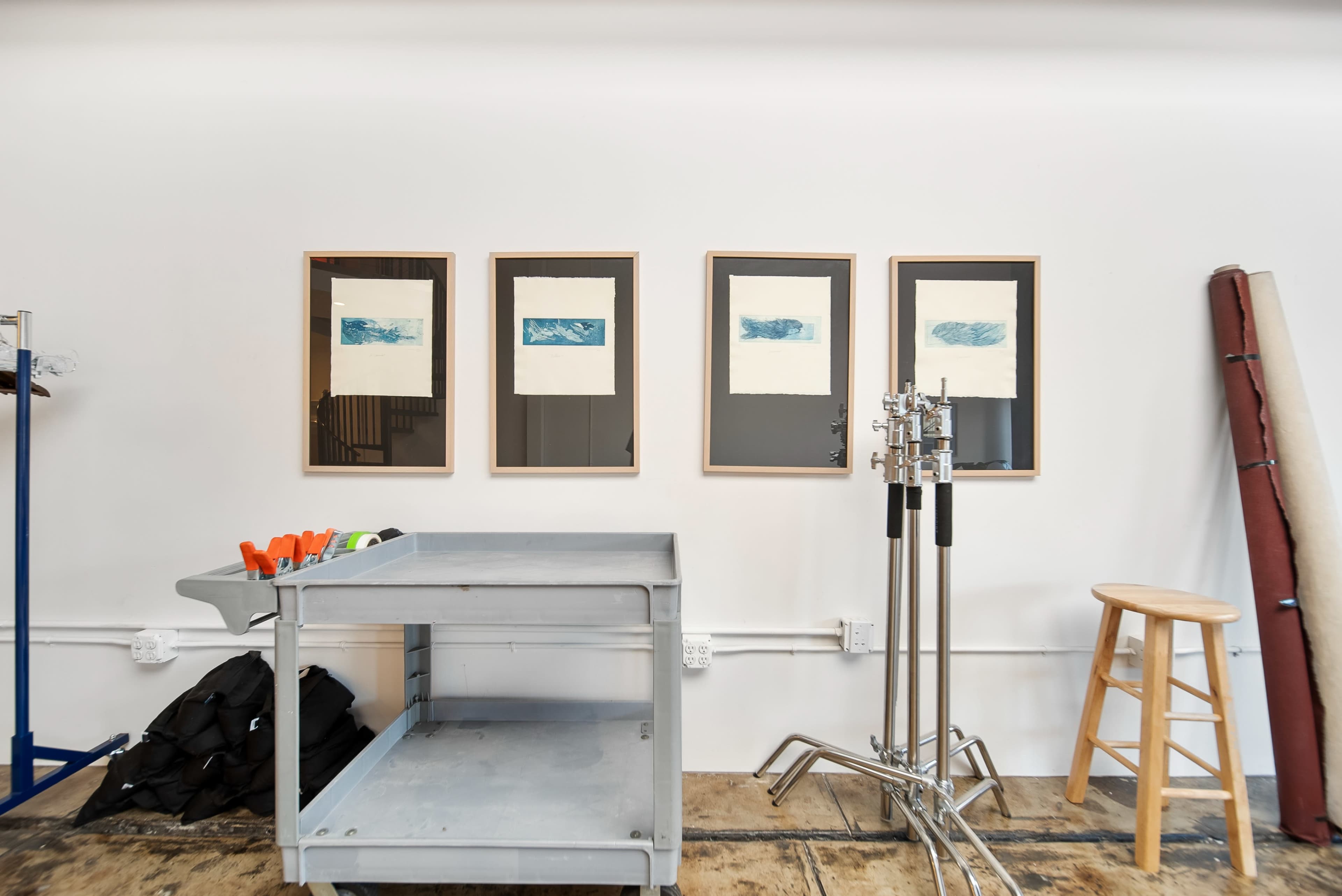 The image shows a well-lit interior space with four framed artworks on the wall and a metal cart in the foreground, accompanied by studio equipment and a wooden stool.