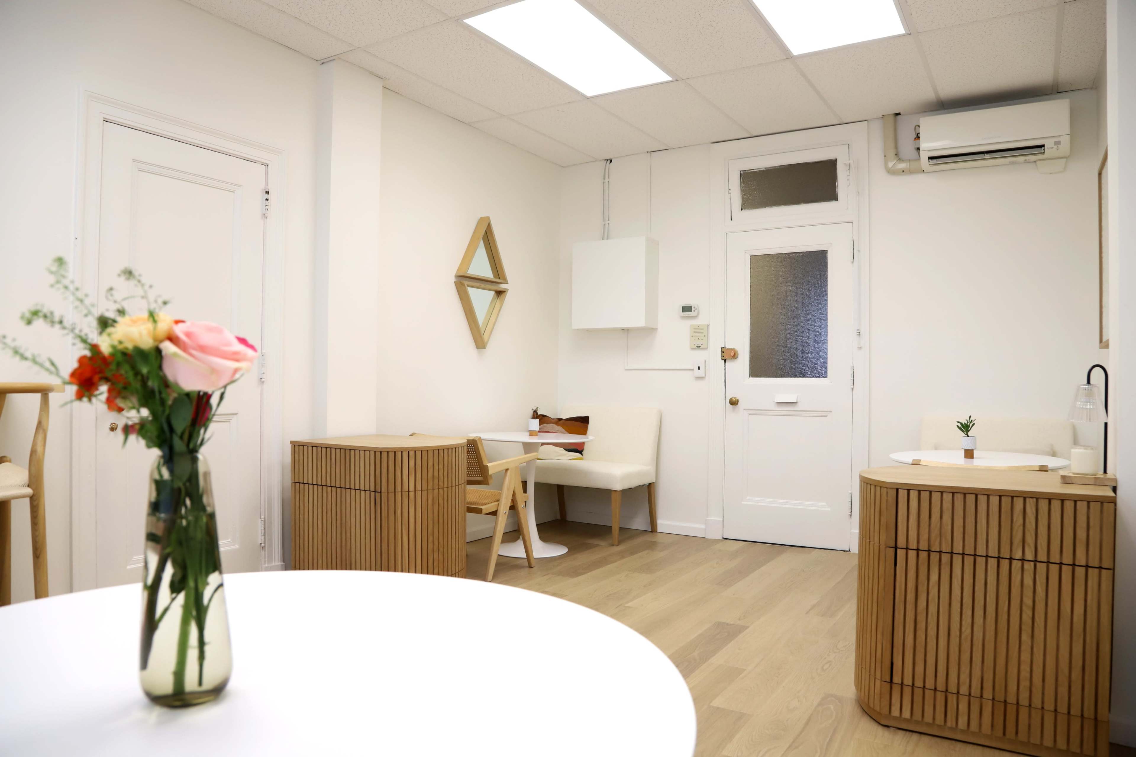 The image shows a bright, minimalist room with light-colored wood furniture, including two square tables, a vase of flowers on one table, and a small mirror on the wall.