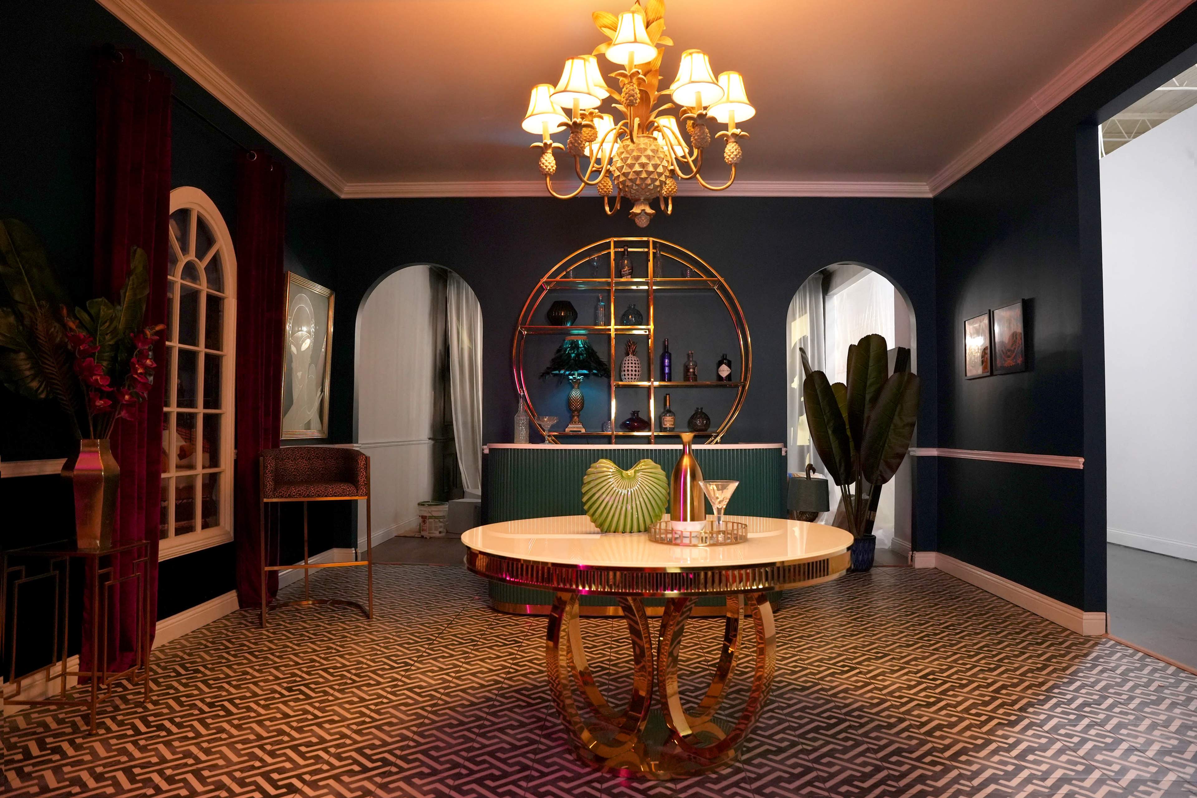 The image shows a stylish room featuring a round table with a gold base, a chandelier, and a bar area decorated with bottles and plants.