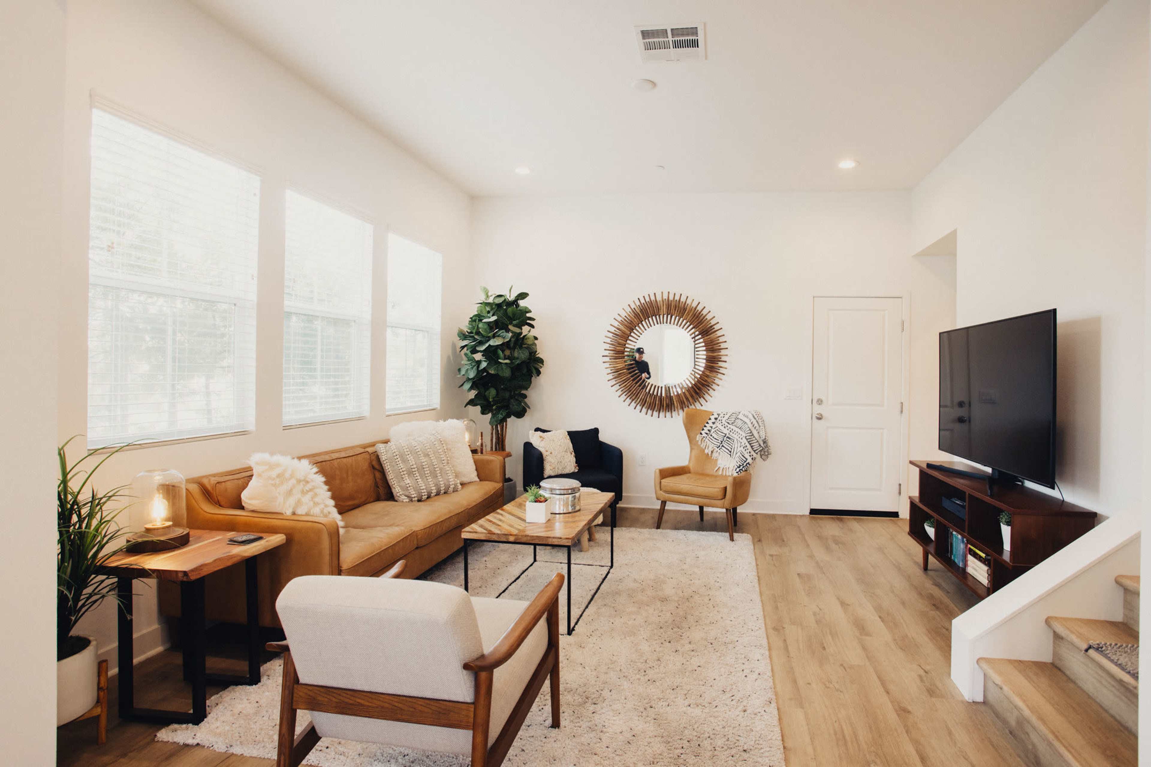 The living room features a tan sofa and armchair, a coffee table, a large mirror, a television, and a potted plant near the window.