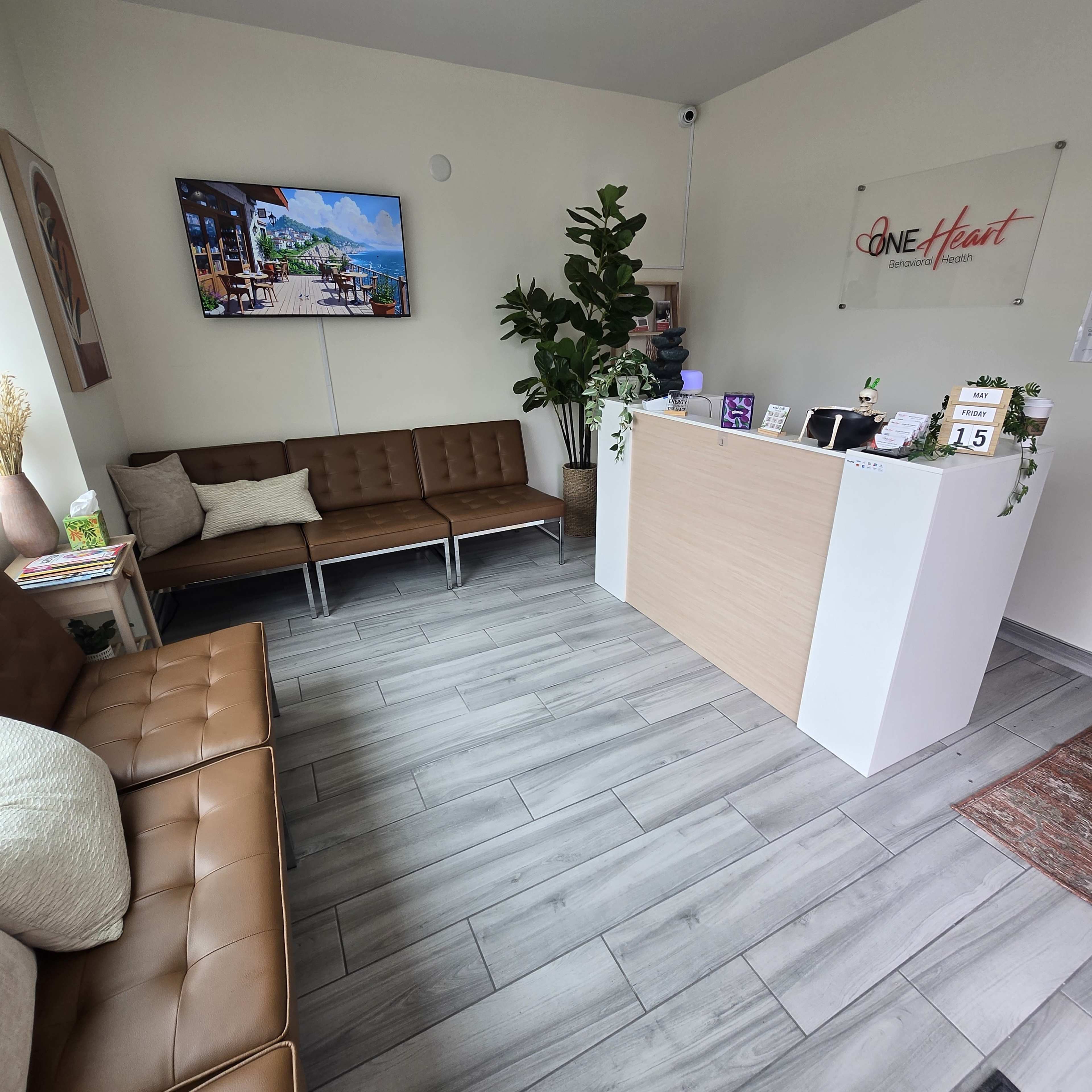 The image shows a bright reception area with modern seating on the left, a sleek front desk on the right, and a television displaying scenic artwork on a wall.