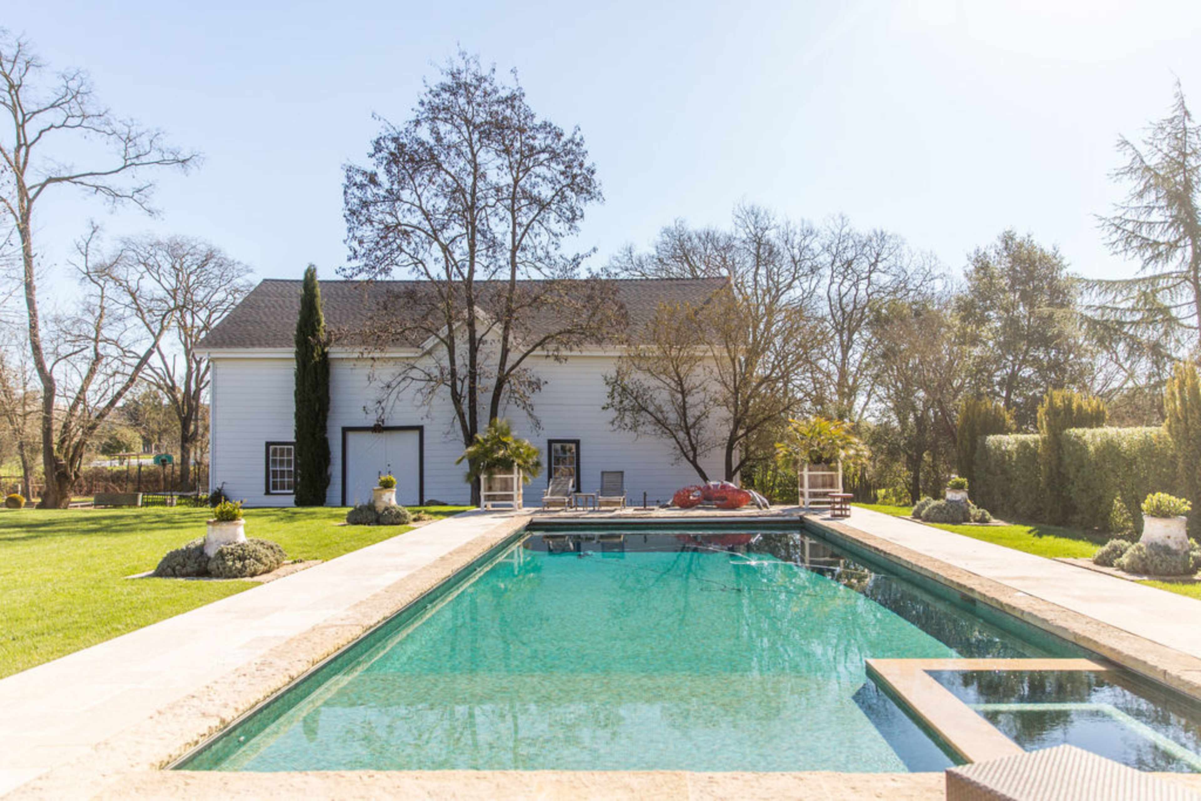A clear rectangular swimming pool set in a garden with a white house and trees in the background.