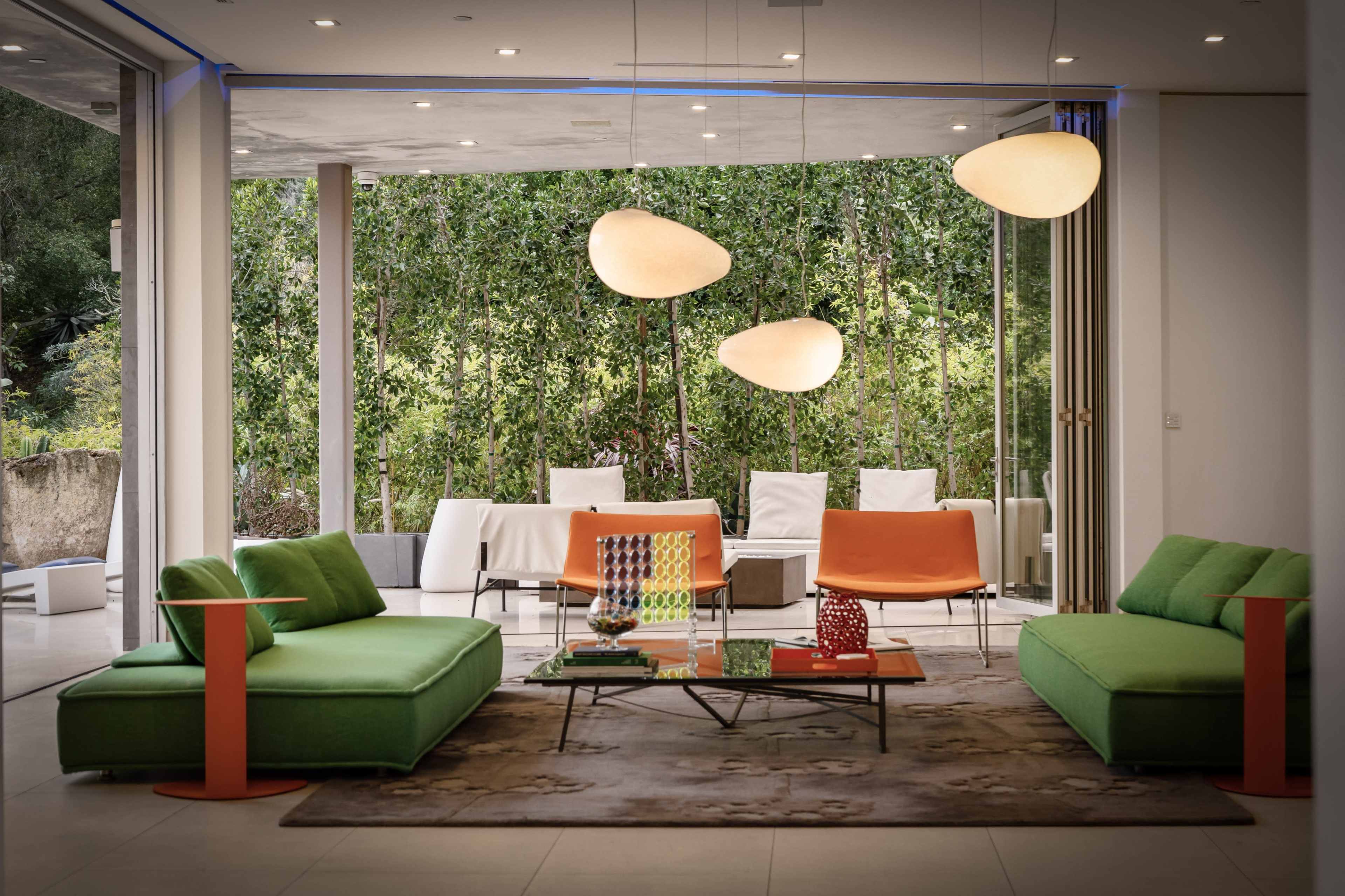 A modern living room features green sofas, orange chairs, and large windows that open to a garden view.