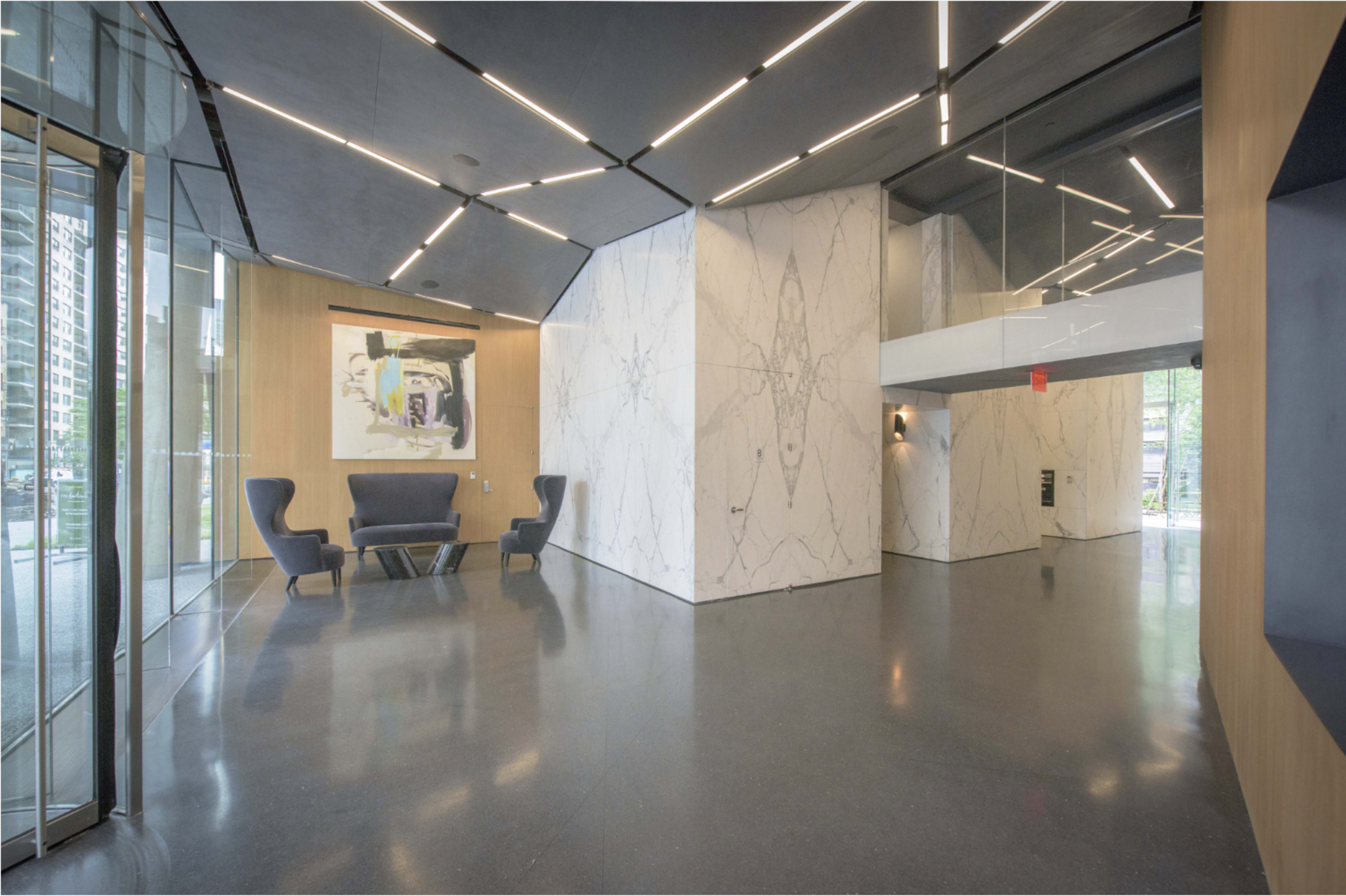 The image shows a modern lobby with sleek walls featuring marble accents, a pair of blue chairs, and large windows letting in natural light.