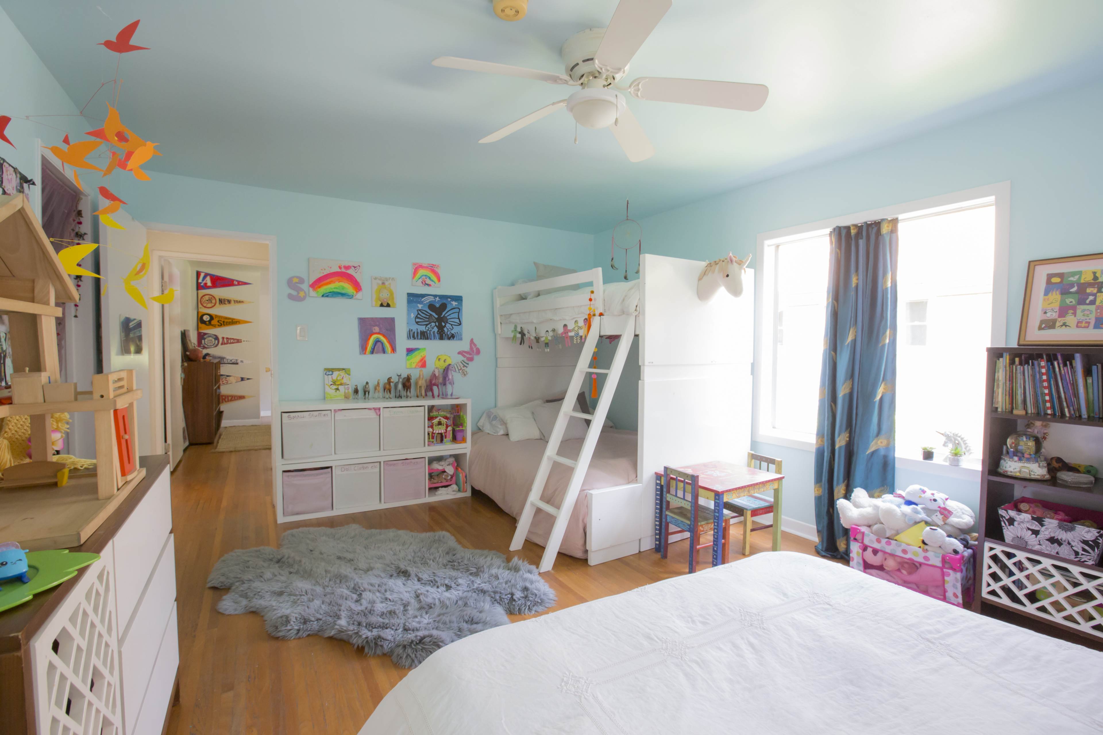 The image shows a light blue children's bedroom with a loft bed, a small table and chairs, and colorful decorations on the walls.