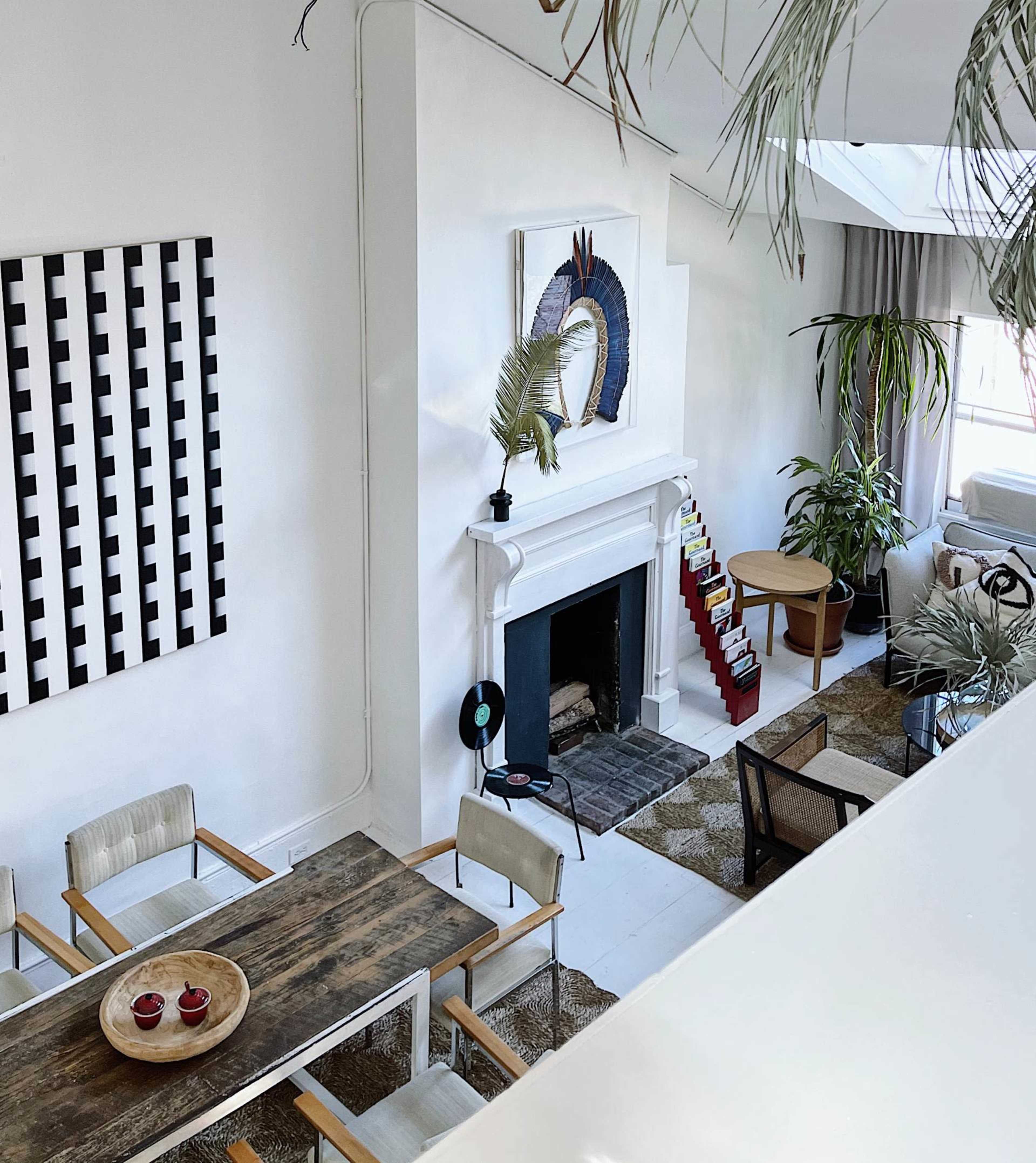 A modern living room with a white fireplace, a dining table, and large plants, accented by a geometric wall art piece.