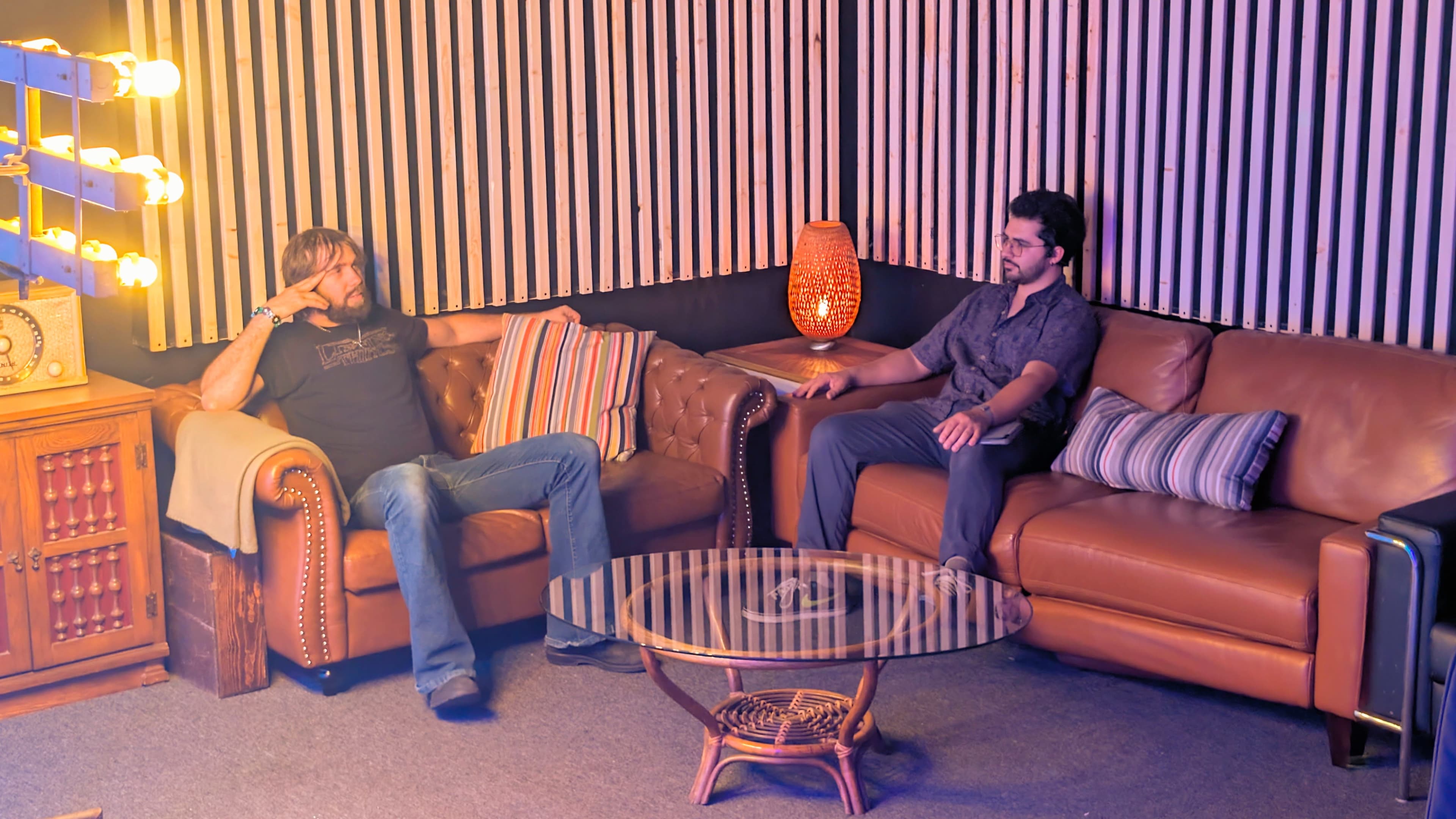 Two men are seated on a pair of leather sofas in a warmly lit room, with decorative lighting and a glass coffee table between them.