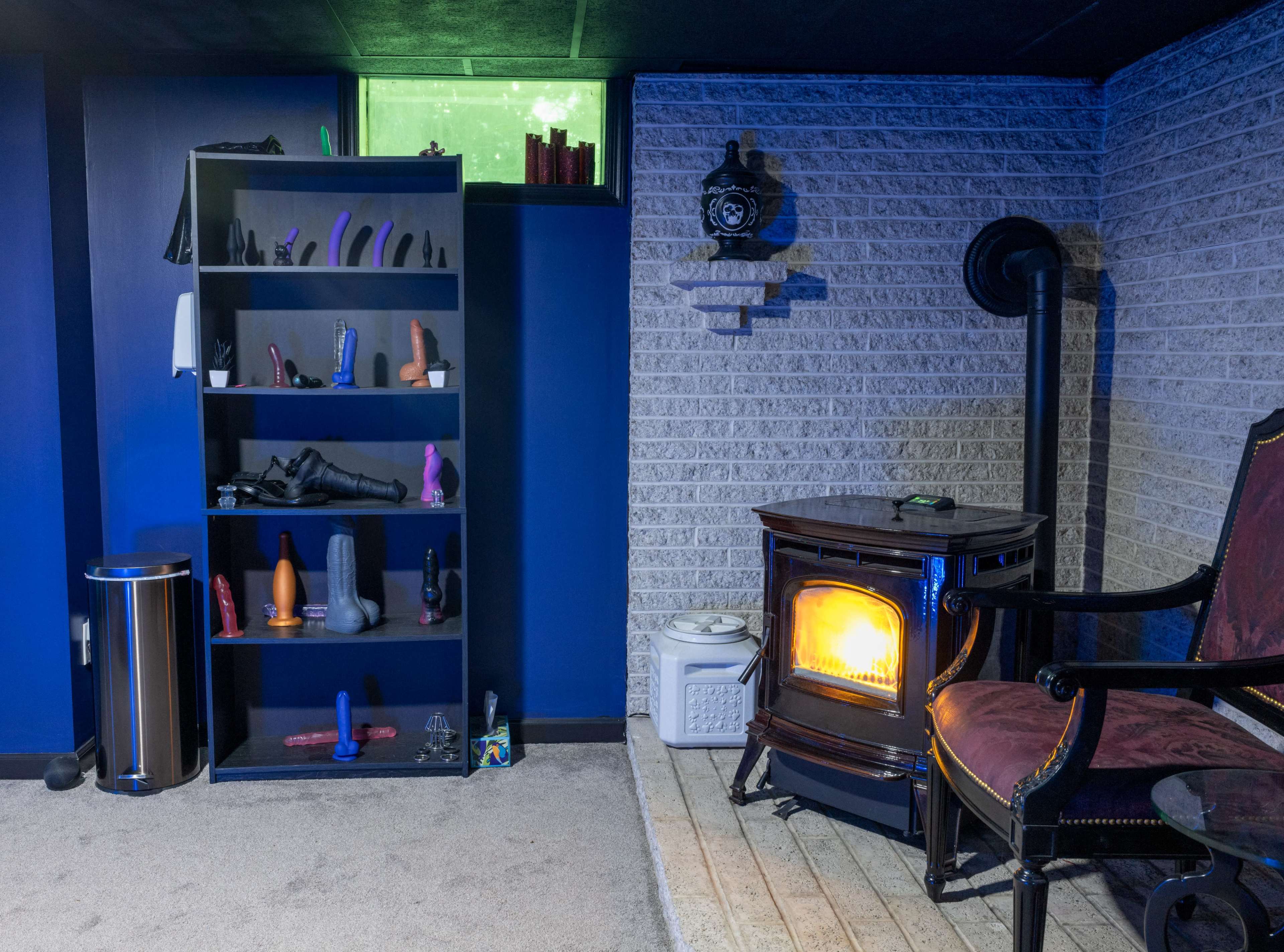 The image shows a dimly lit basement room featuring a black shelving unit filled with various items, a comfortable chair beside a heating stove, and a window allowing some light to enter.