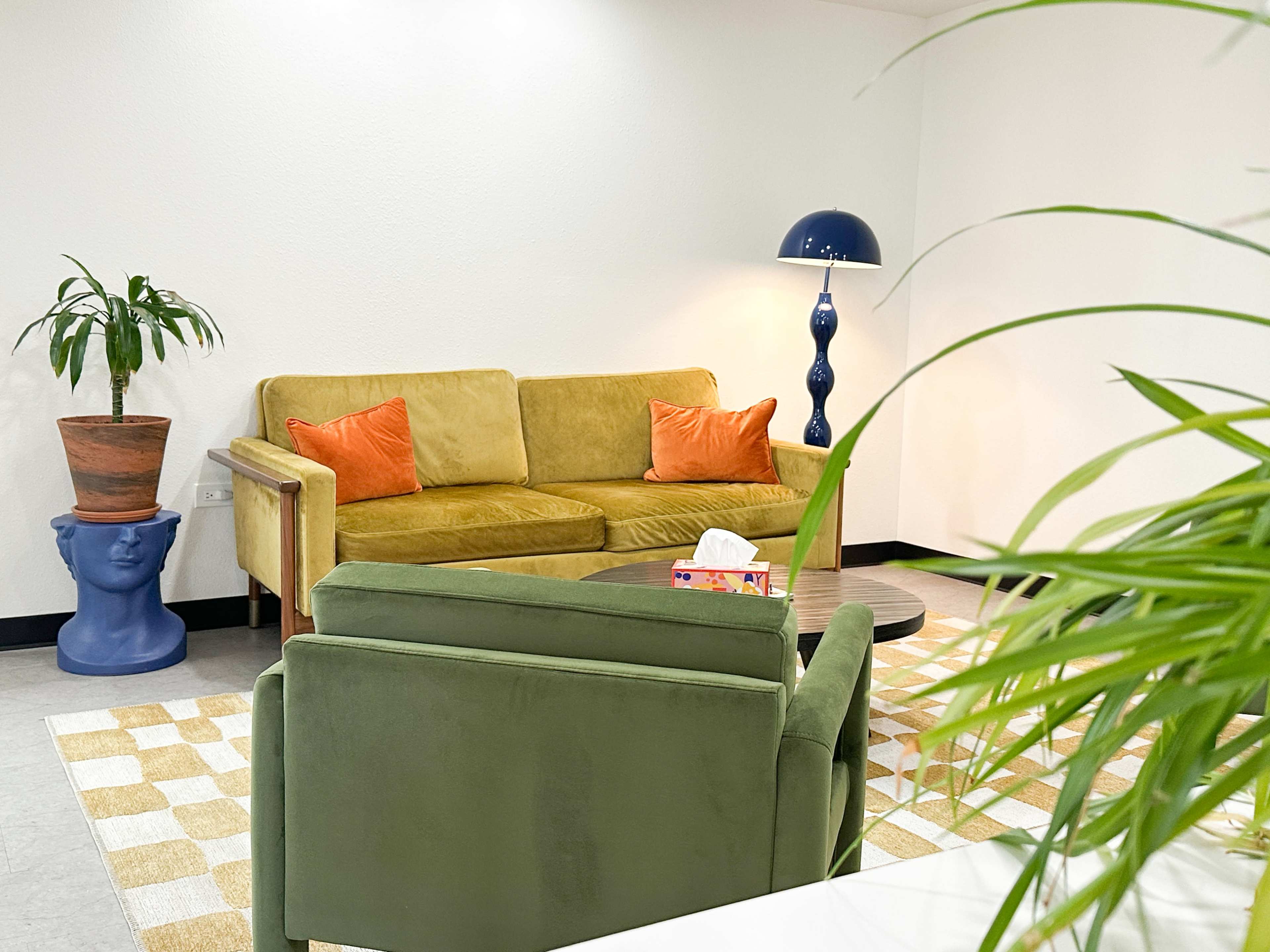 The image shows a modern lounge area featuring a yellow sofa with orange cushions, a blue-themed lamp, and a green armchair surrounded by decorative plants.