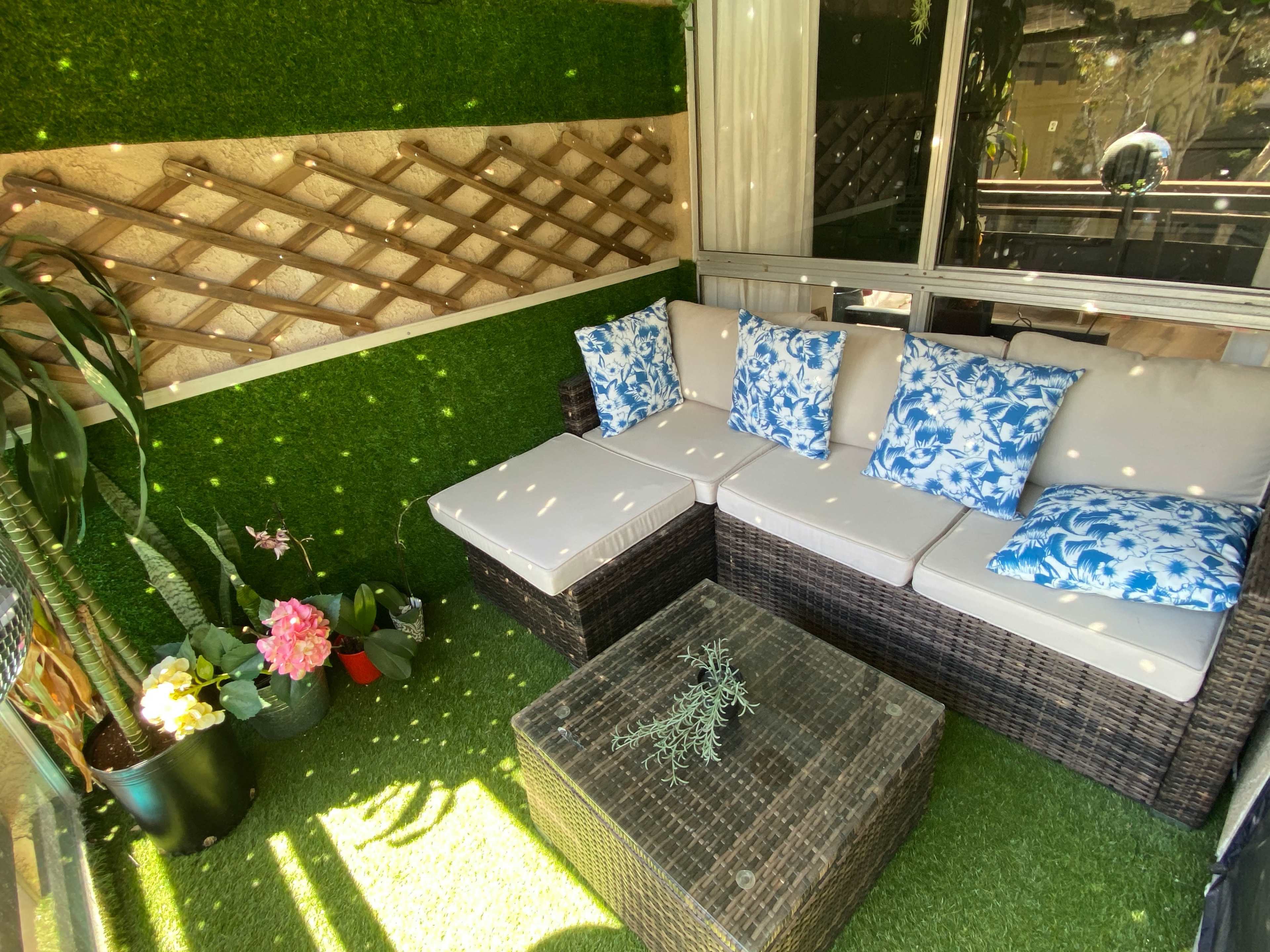 A cozy outdoor seating area features a wicker sofa with blue floral cushions, surrounded by artificial grass and potted plants.