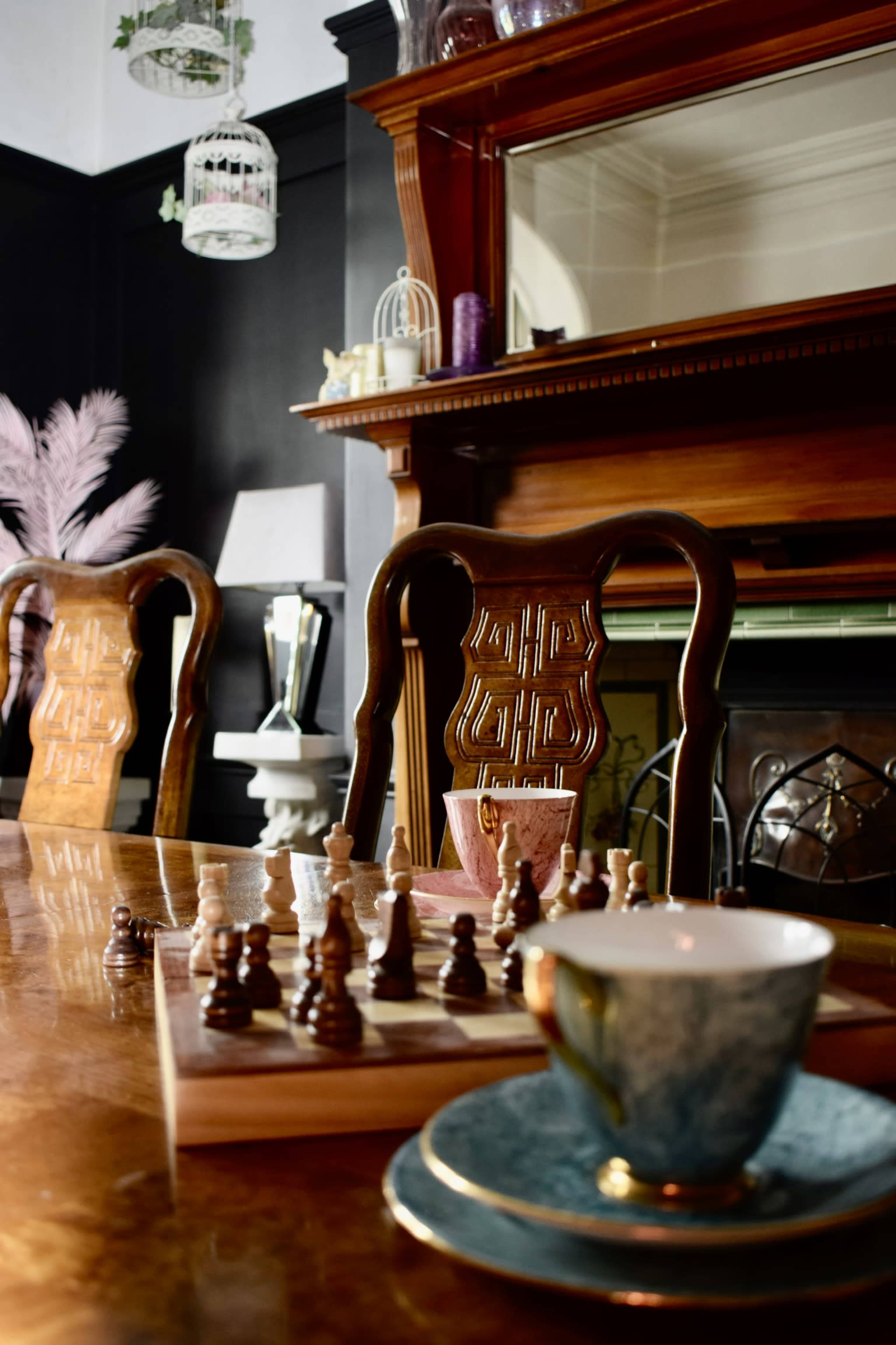 A wooden table displays a chess set in front of an ornate mirror and a fireplace, surrounded by three decorative chairs and a tea cup on a saucer.