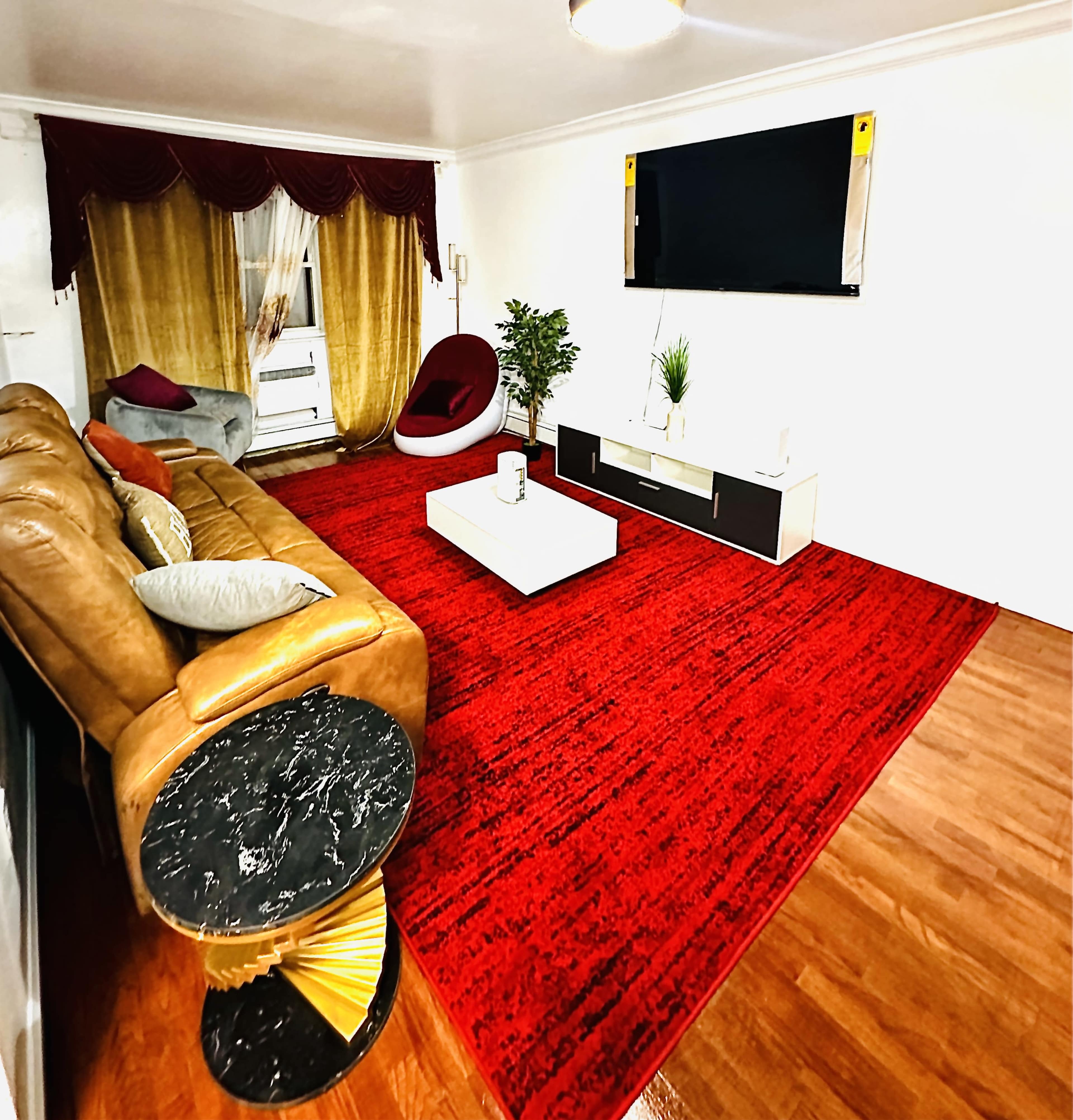 A modern living room featuring a brown leather sofa, a large red area rug, a white coffee table, and a wall-mounted television.