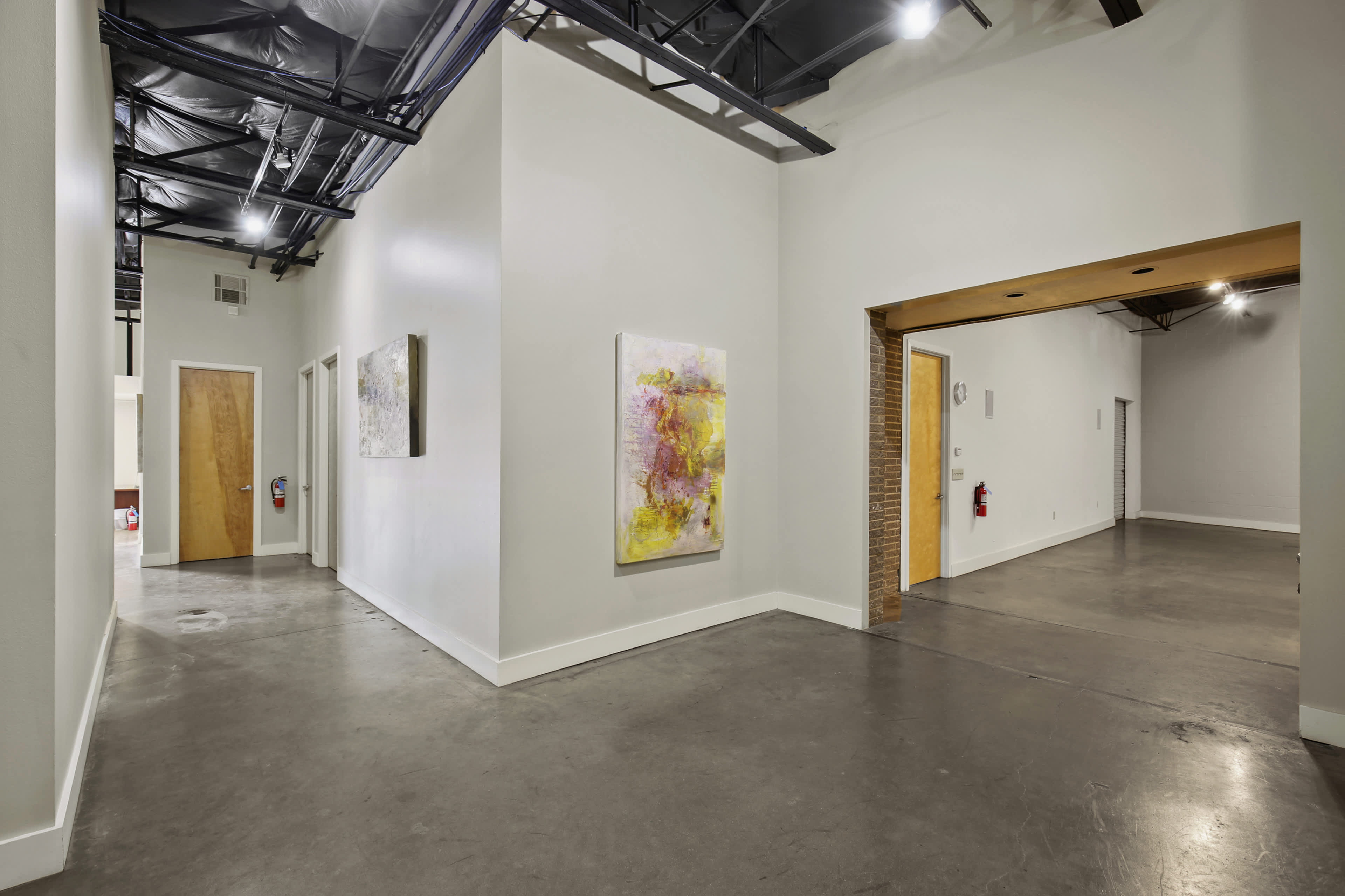 The image shows a spacious, modern hallway featuring white walls, concrete flooring, and framed artwork on the walls, leading to an open doorway.