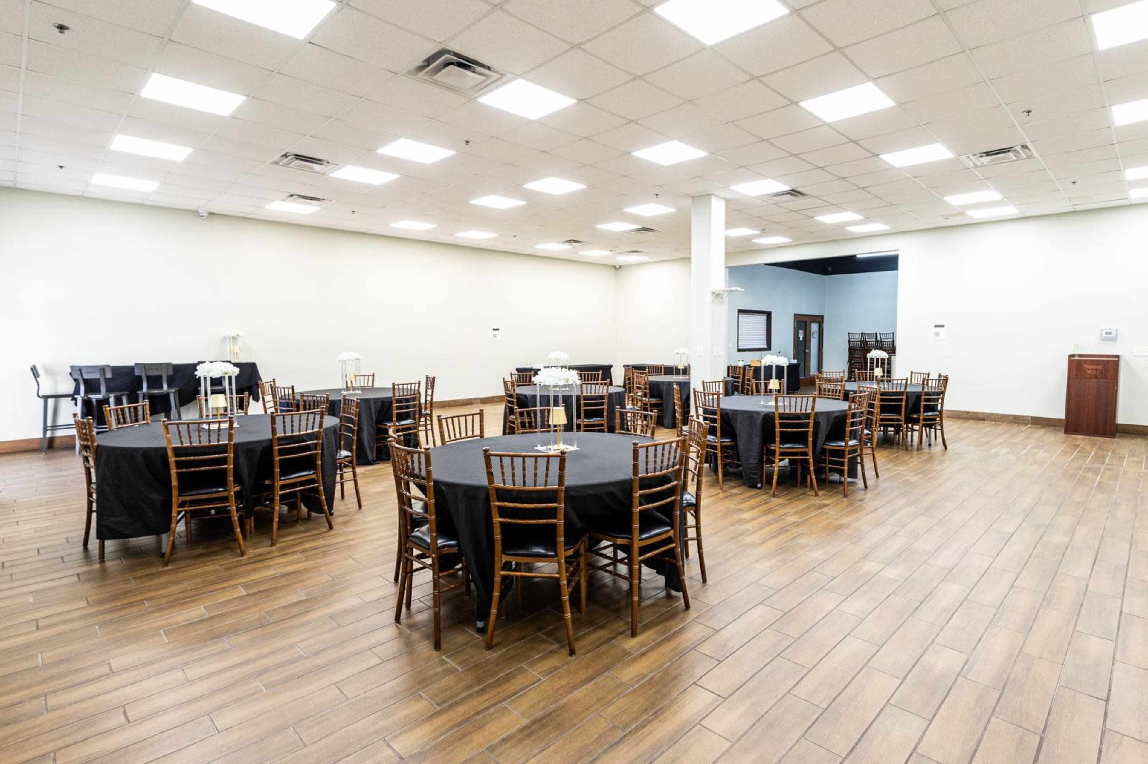 The image shows a spacious event room with several round tables covered in black cloth and surrounded by wooden chairs.