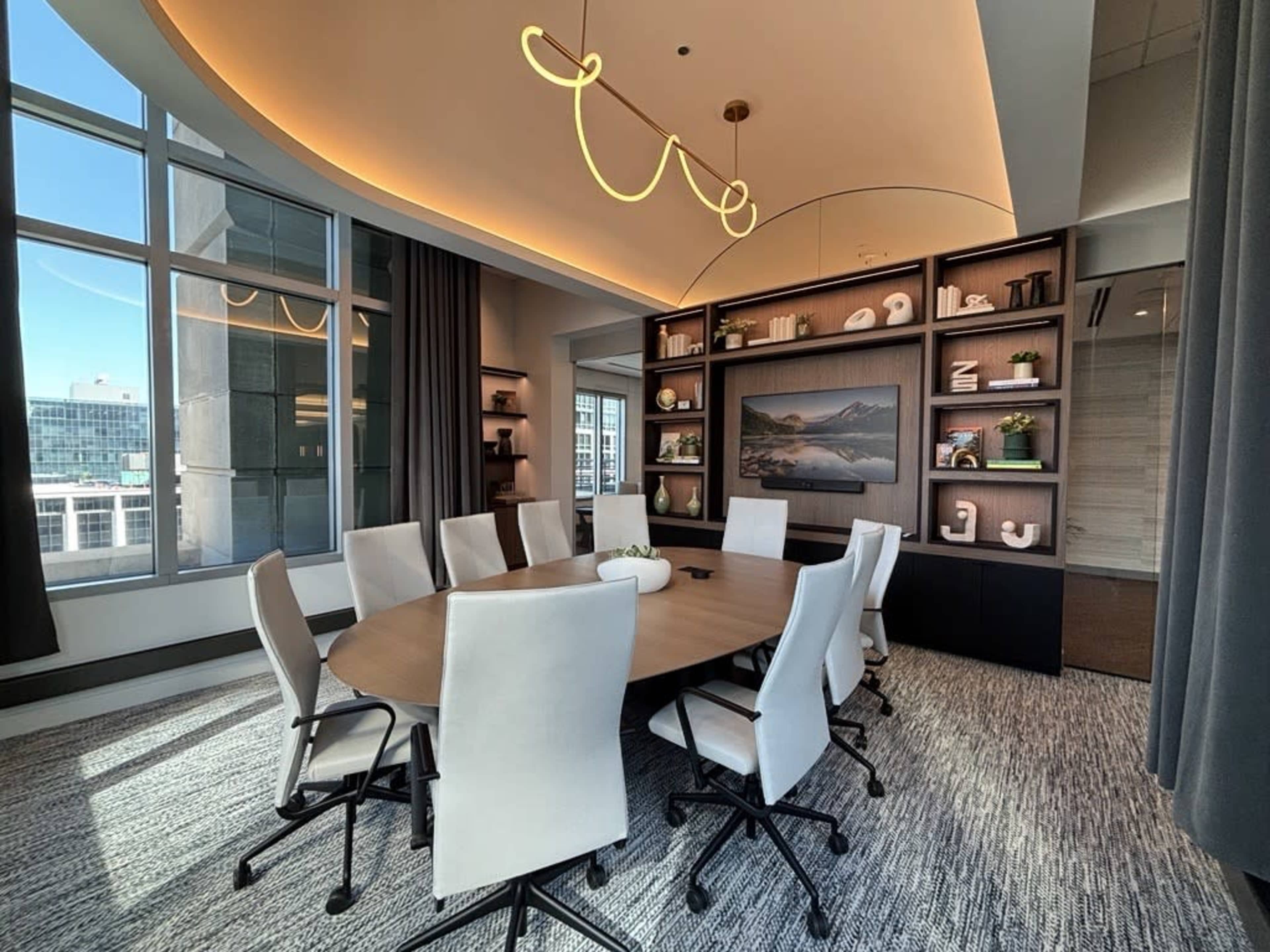 A modern conference room features a large round table surrounded by white chairs, with a wall display of decorative items and a view through large windows.