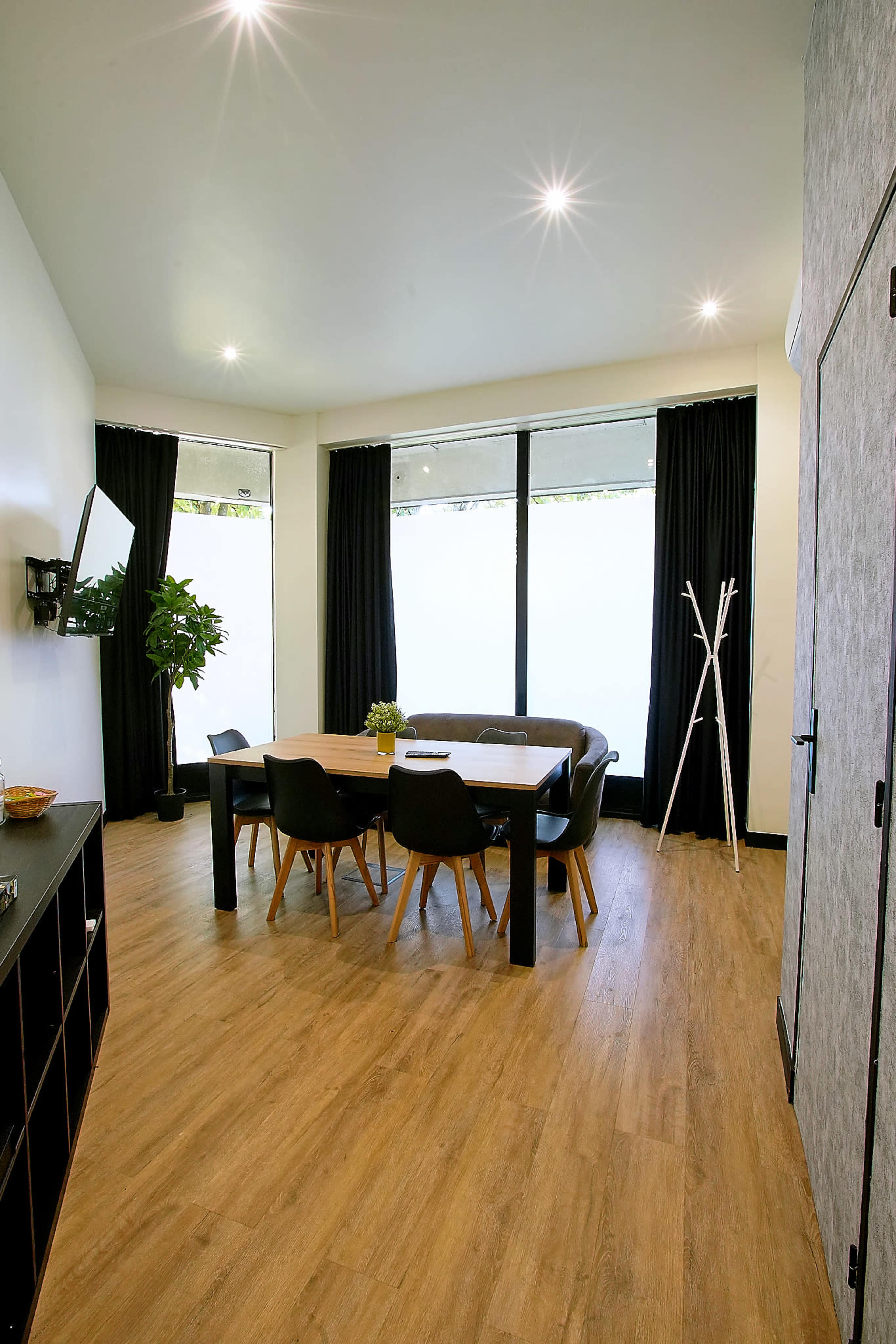 The image shows a modern dining area with a wooden table surrounded by black chairs near large windows covered with dark curtains.