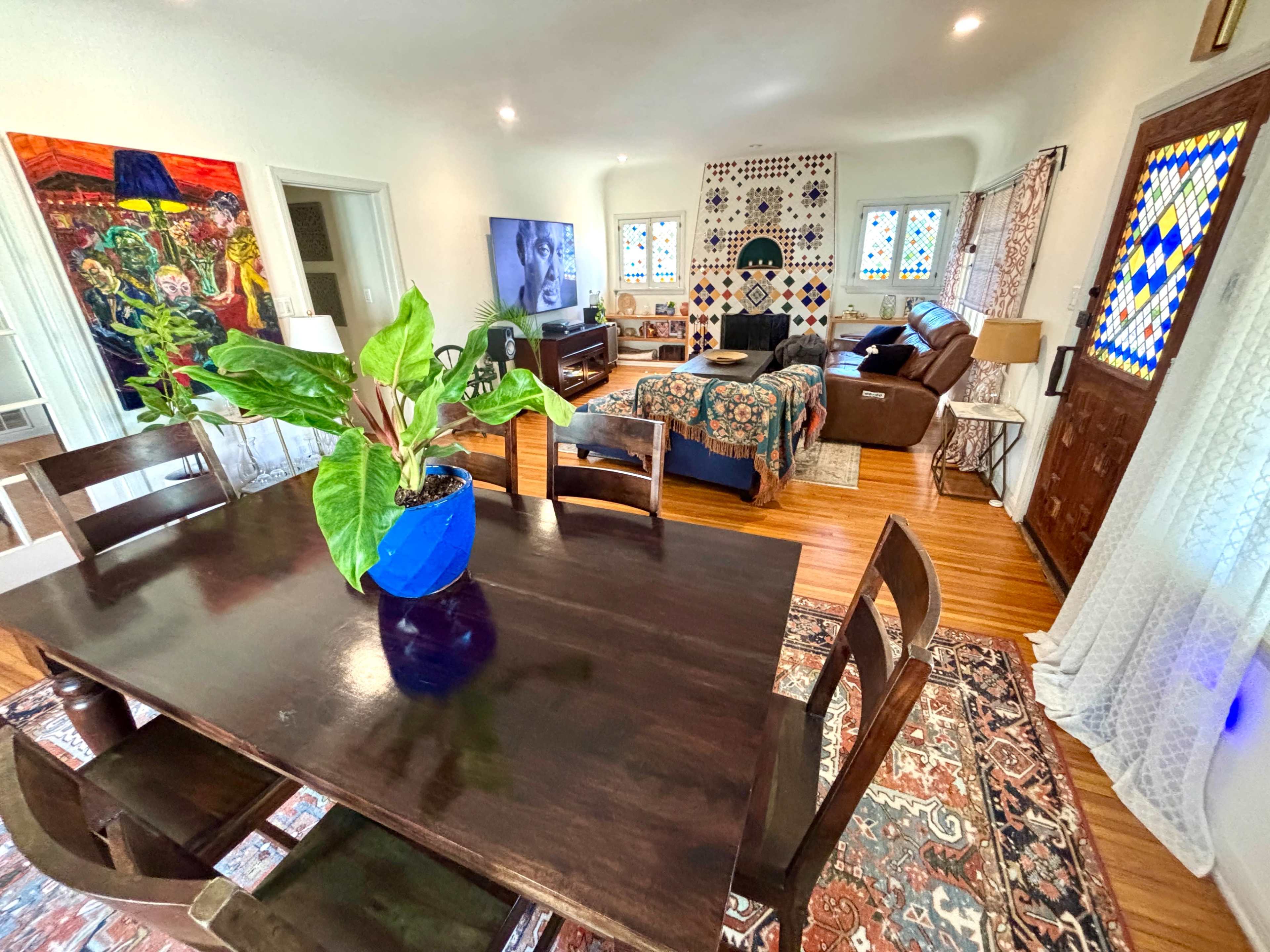 The image shows a well-lit room featuring a dining table with a plant, a living area with a television, and a bedroom area with a colorful bedspread and decorative windows.
