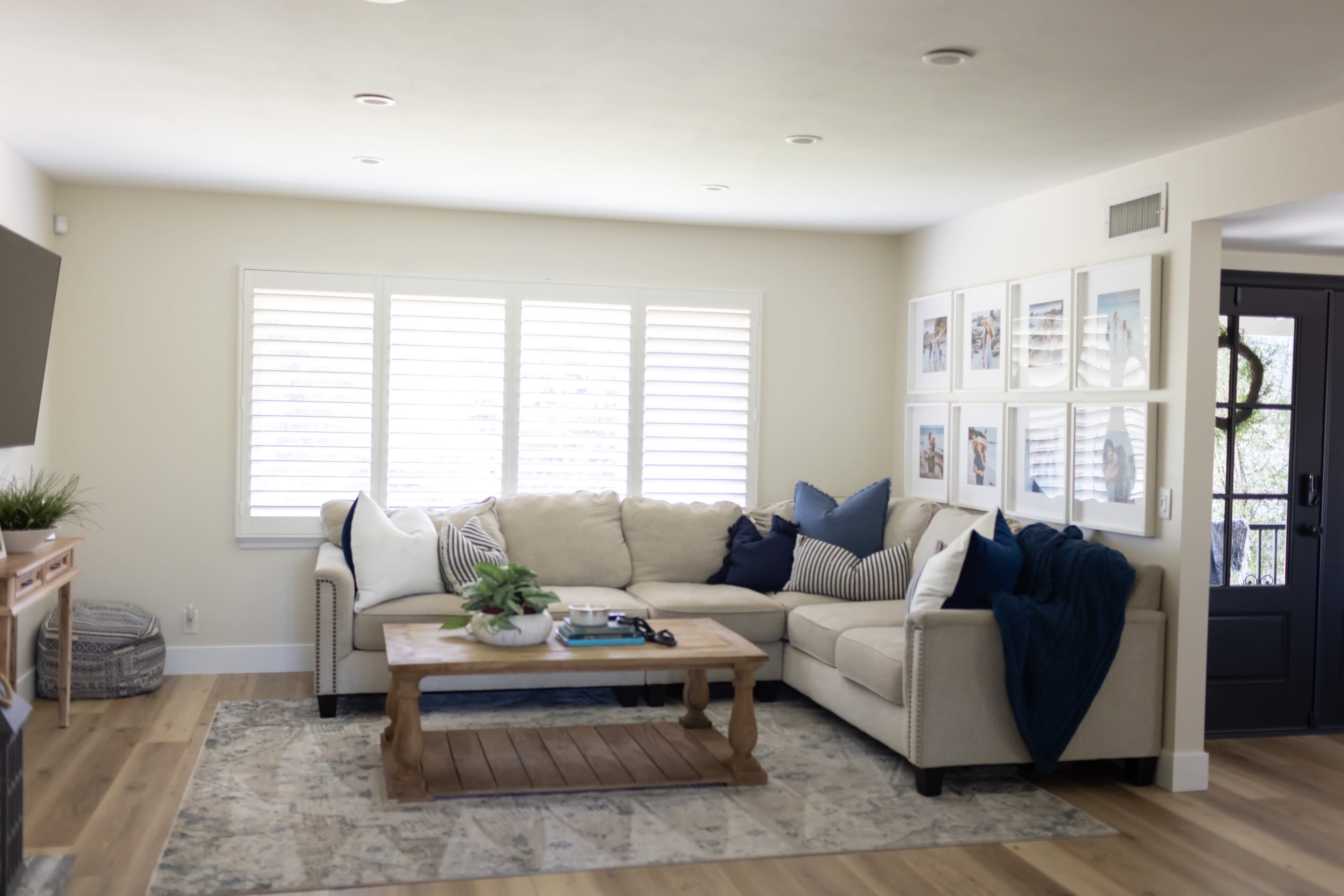 The image shows a clean living room with a beige sectional sofa, a wooden coffee table, and framed pictures on the wall beside a set of shuttered windows.