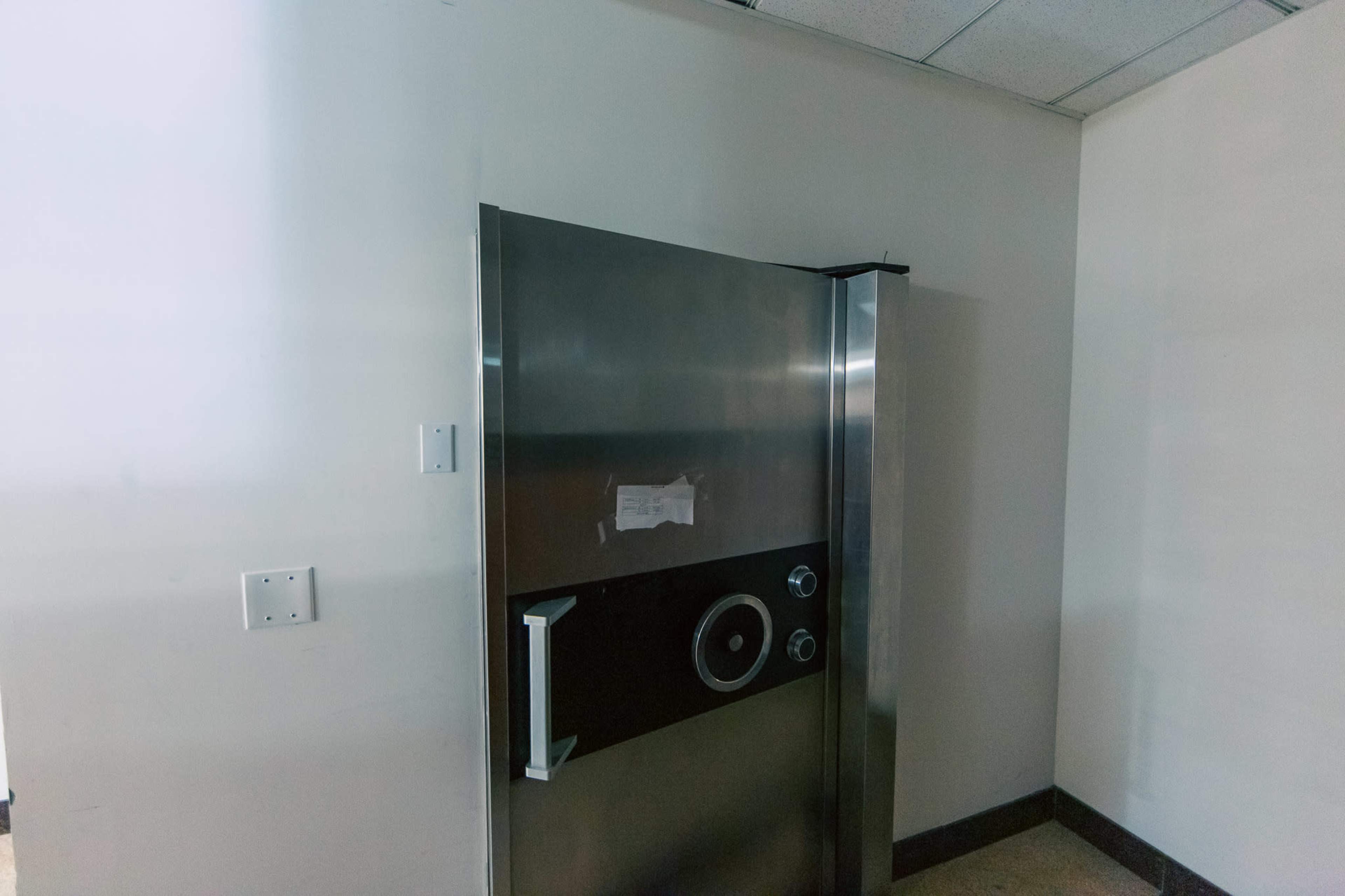 The image shows a large, metallic safe door installed against a plain wall in a room with overhead lighting.