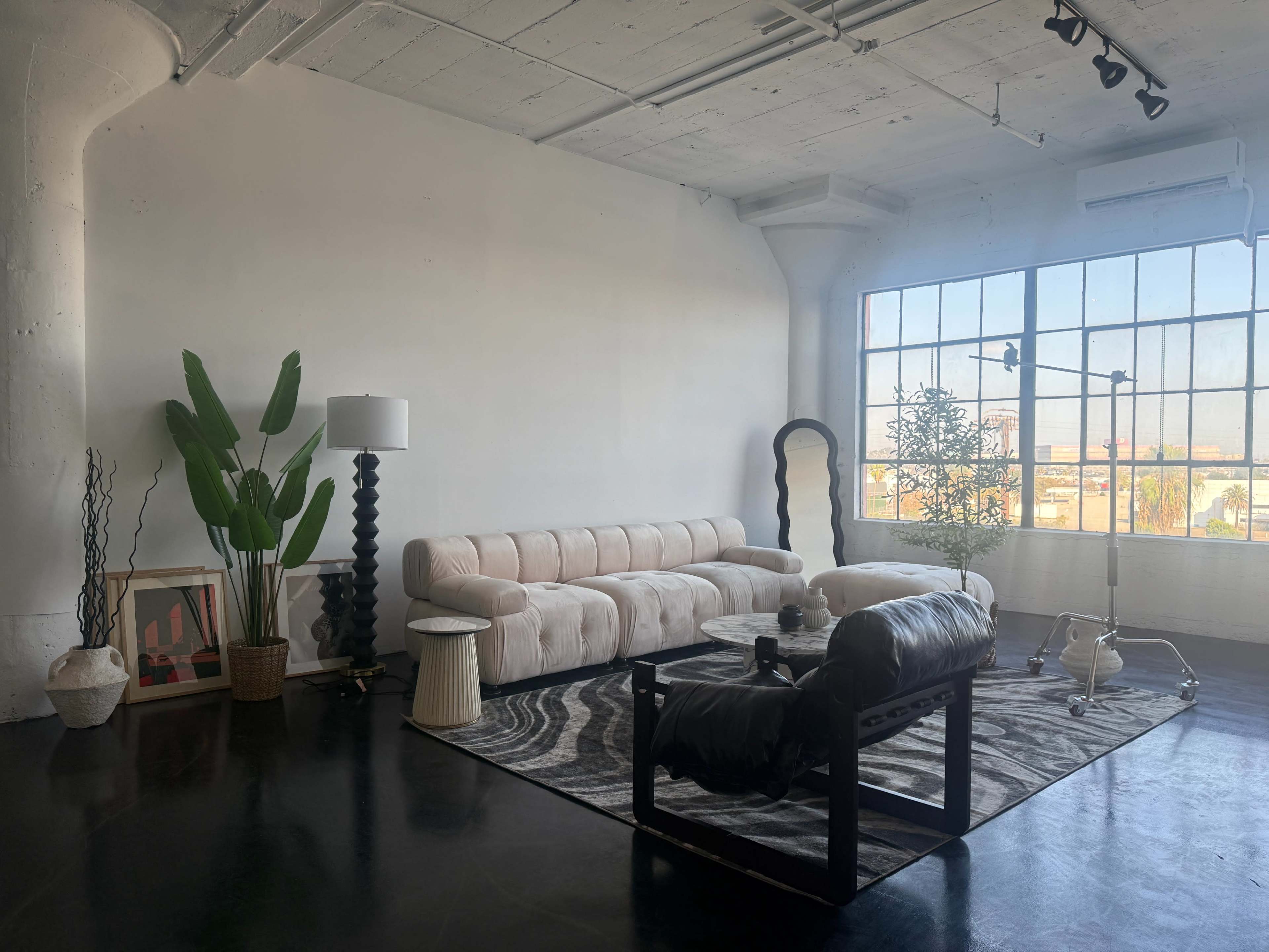 The image shows a modern interior living space with a large window, a beige couch, a black accent chair, a round coffee table, and decorative plants.