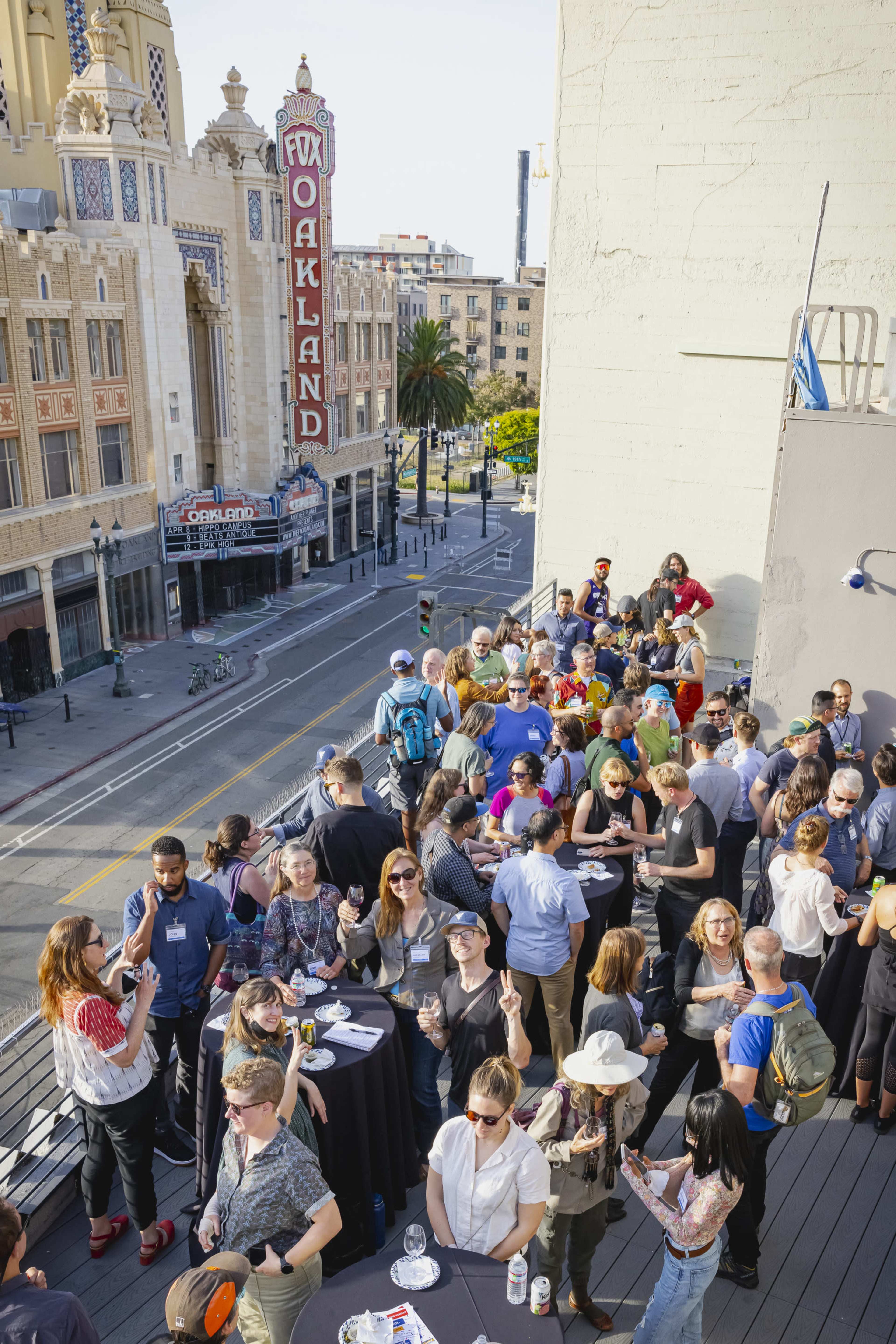 A large group of people gathers on a rooftop terrace overlooking the Fox Oakland Theatre and the street below.