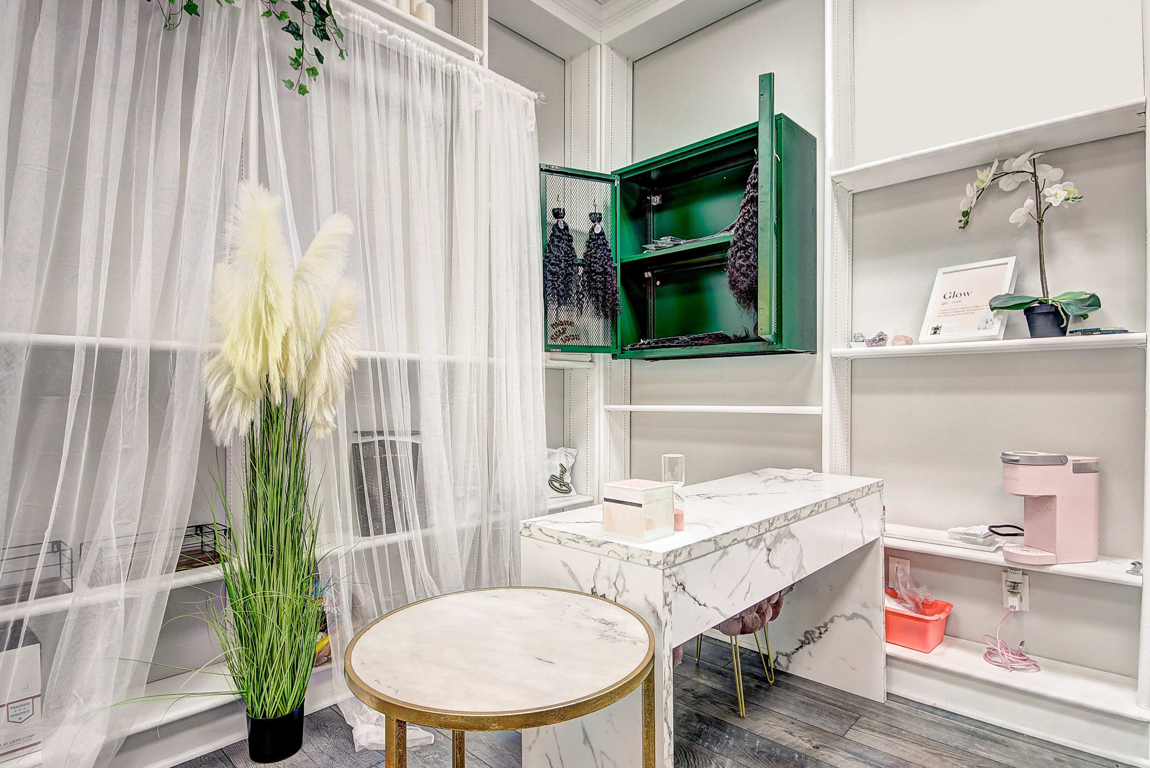 A modern room with a white sheer curtain, a marble-top desk, a round gold-legged table, a green cabinet, and decorative plants.