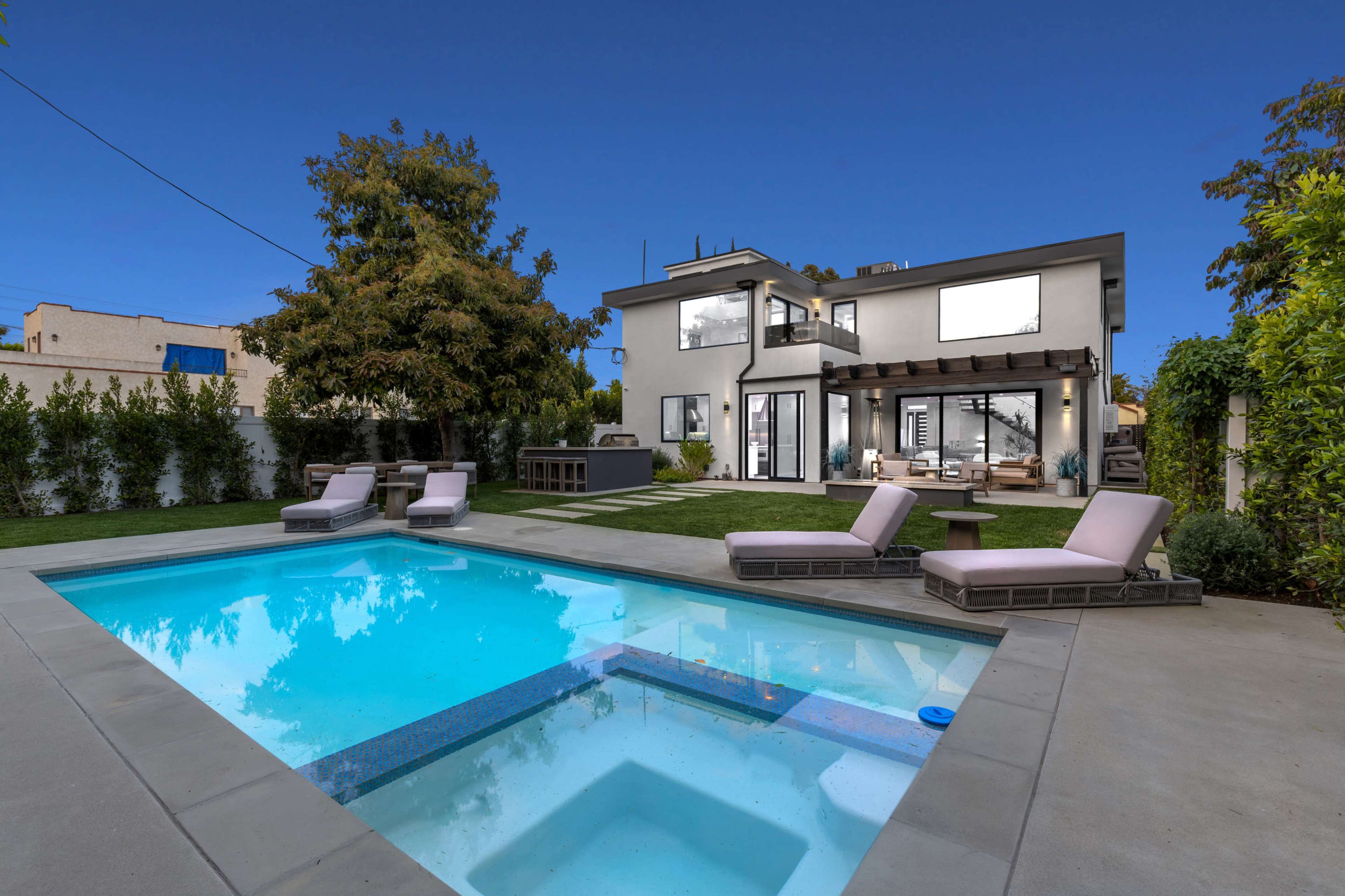The image shows a modern two-story house with a swimming pool and lounge chairs in the backyard, surrounded by lush greenery.