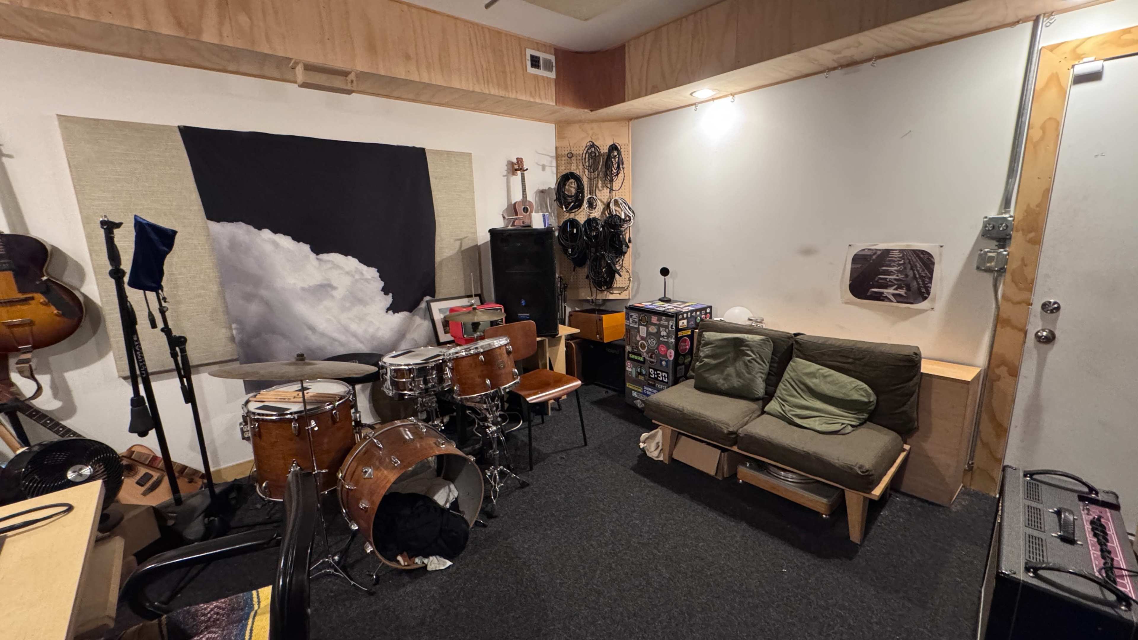 The image shows a cluttered music rehearsal space with a drum set, sofa, and various musical equipment against the walls.