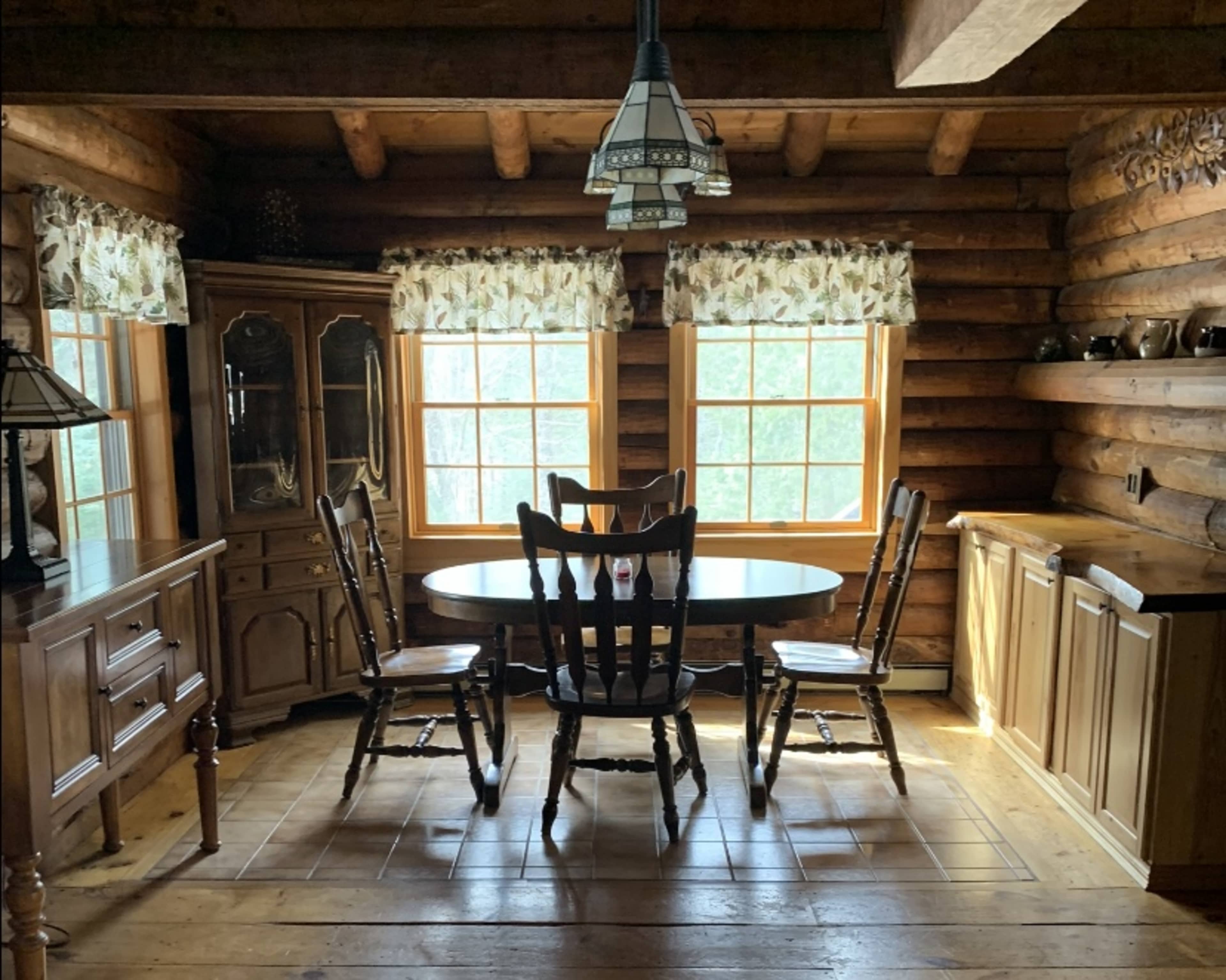 A wooden dining table surrounded by four chairs is situated in a log cabin kitchen with large windows and rustic furniture.