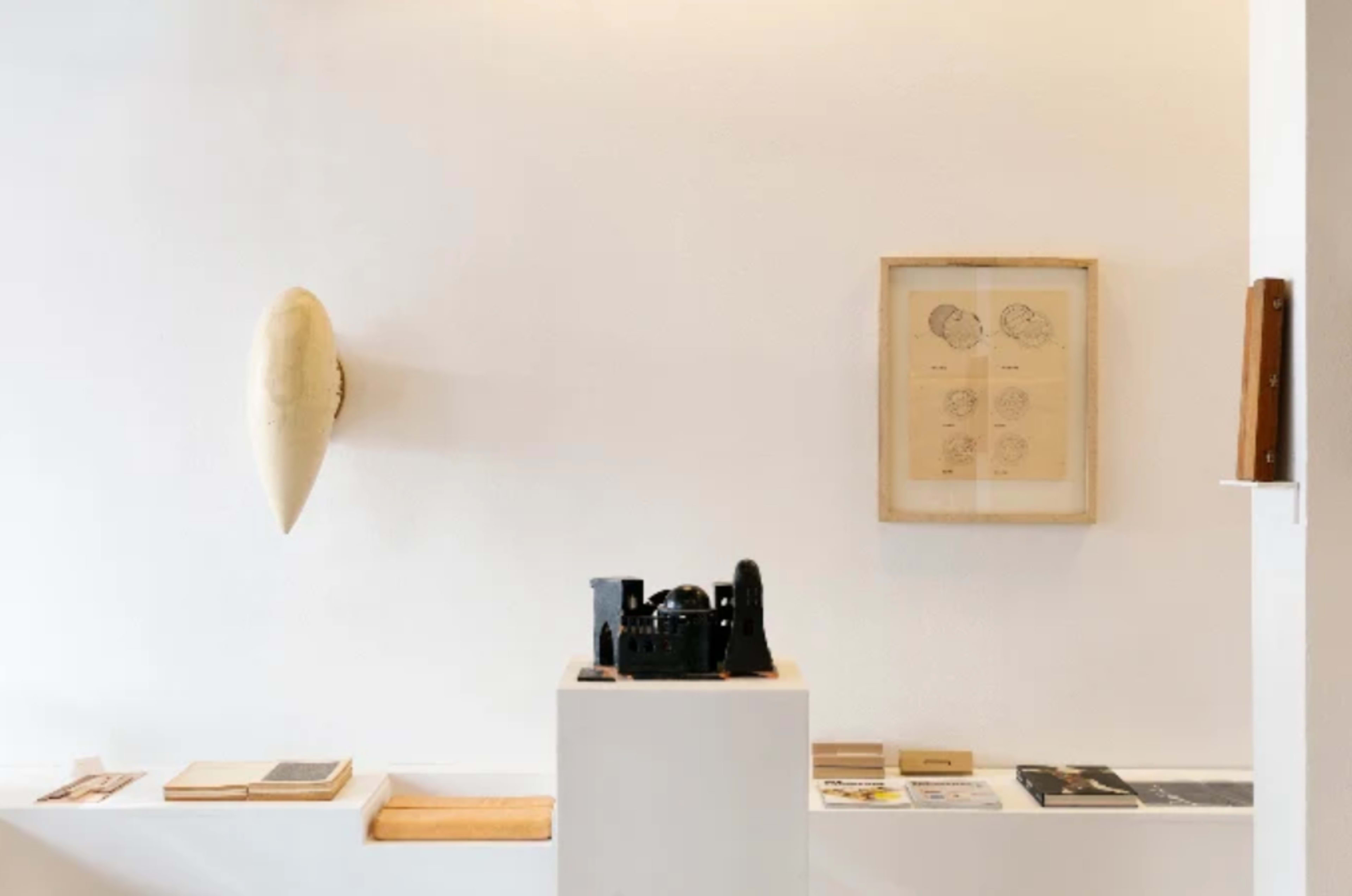 The image shows a minimalist interior featuring a mounted sculpture on the wall, a framed illustration, and a pedestal displaying a black object, along with various books and materials arranged on a ledge.