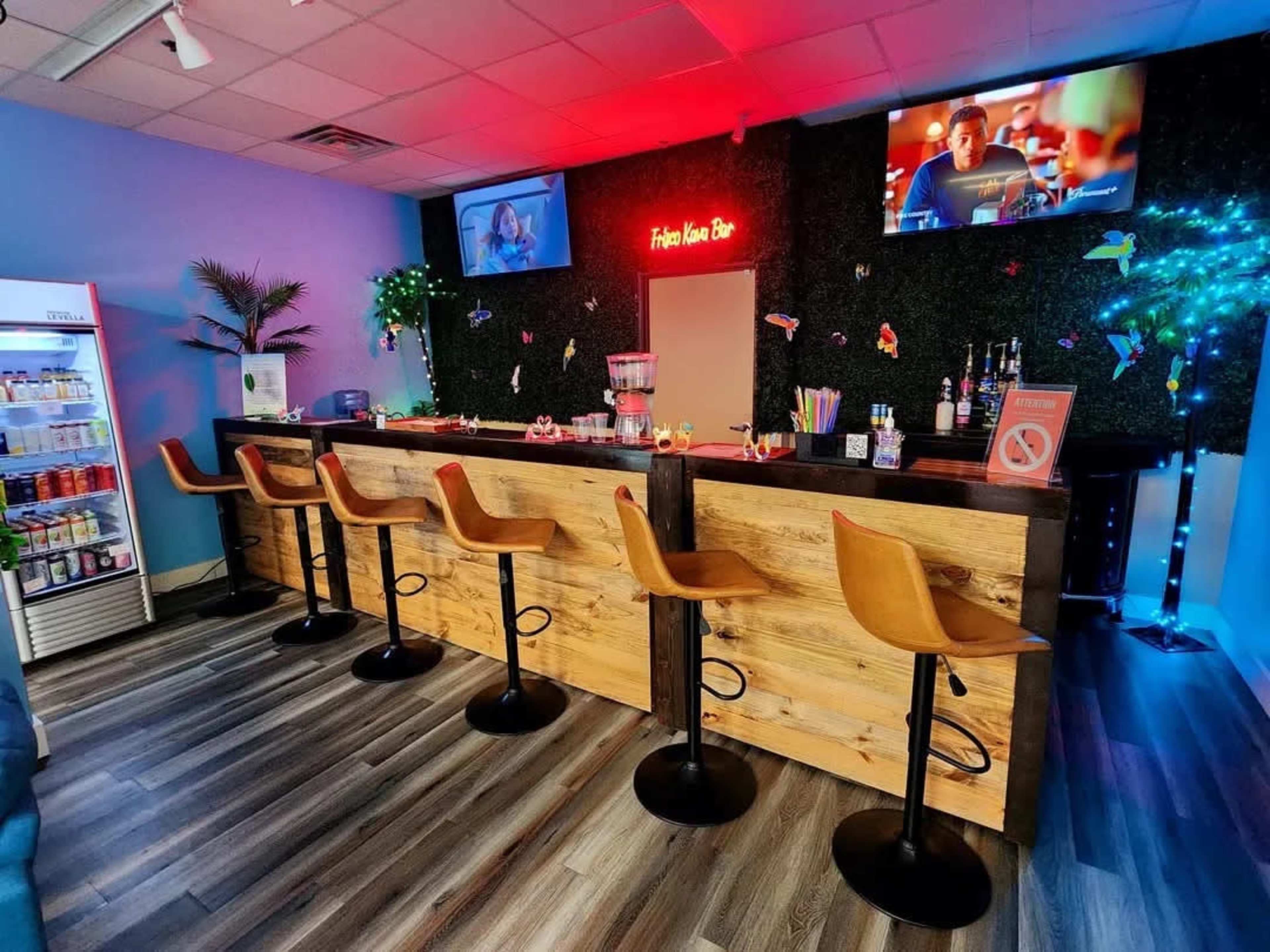 The image shows a colorful bar area with wooden countertops, several high stools, and two televisions mounted on the wall, all under ambient lighting.