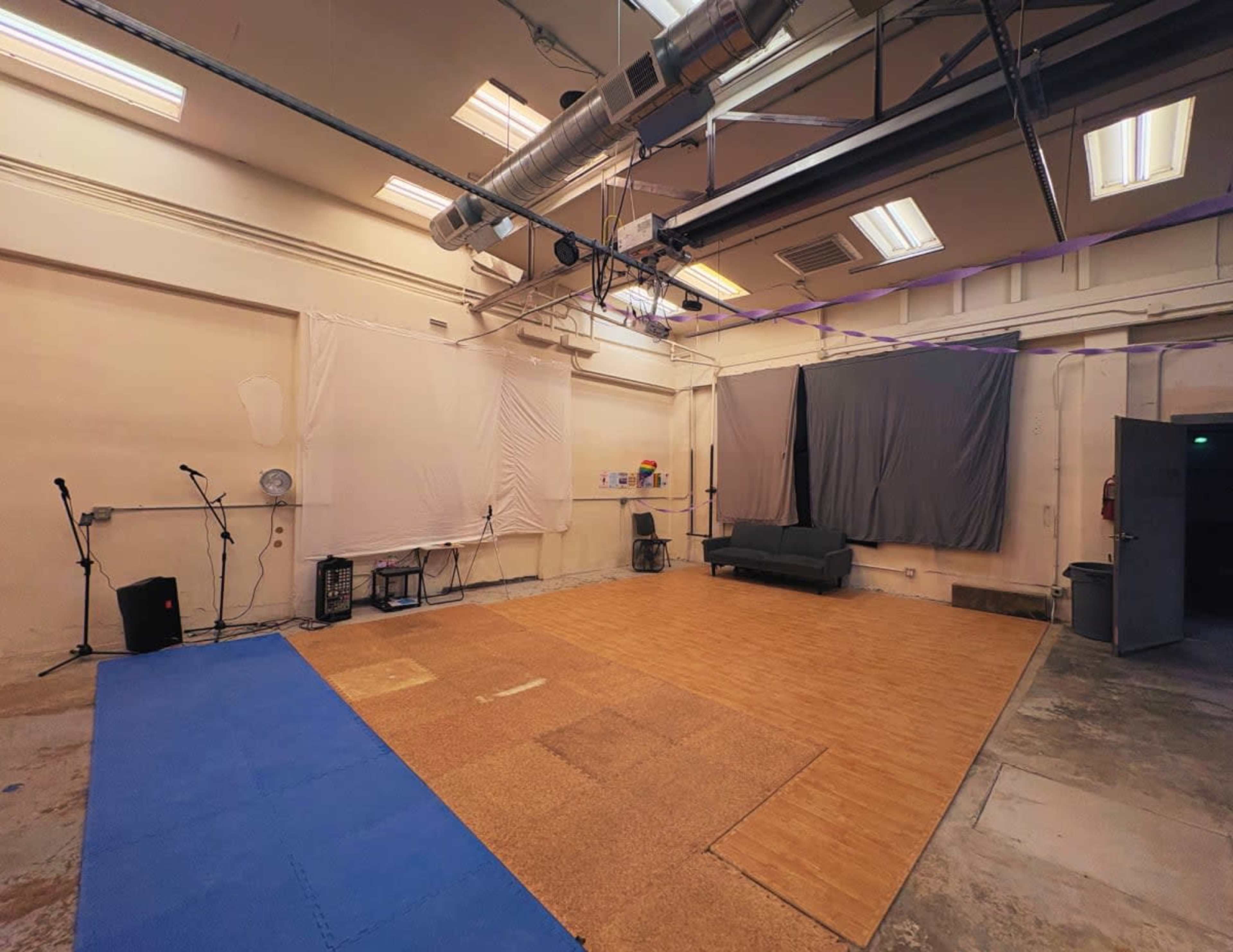 The image shows an empty studio space with a wooden floor, a couch against the wall, and equipment for sound recording or performance set up in the corners.