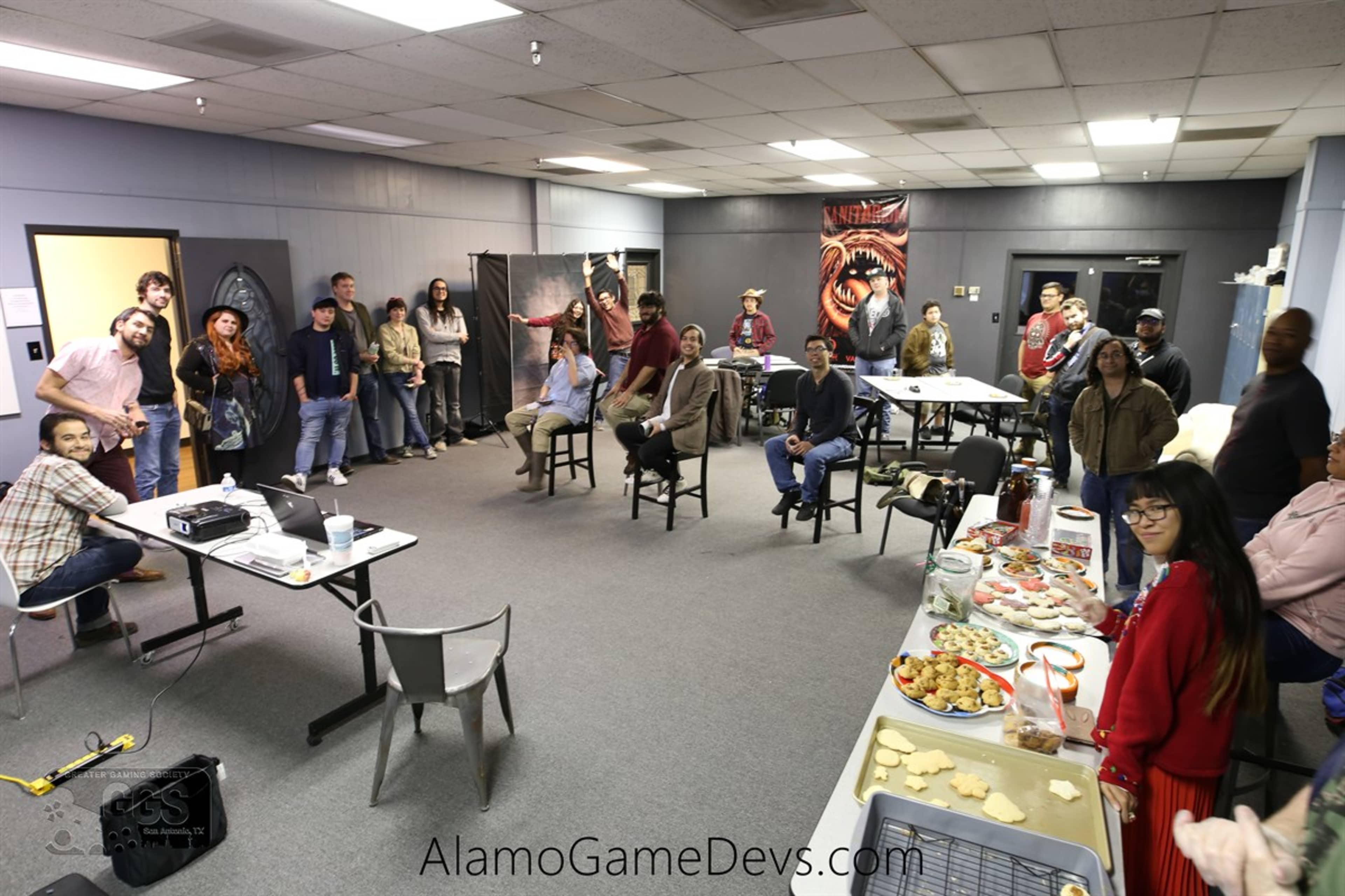 A group of people is gathered in a large room with tables and snacks, engaging in conversation while some are seated and others are standing.