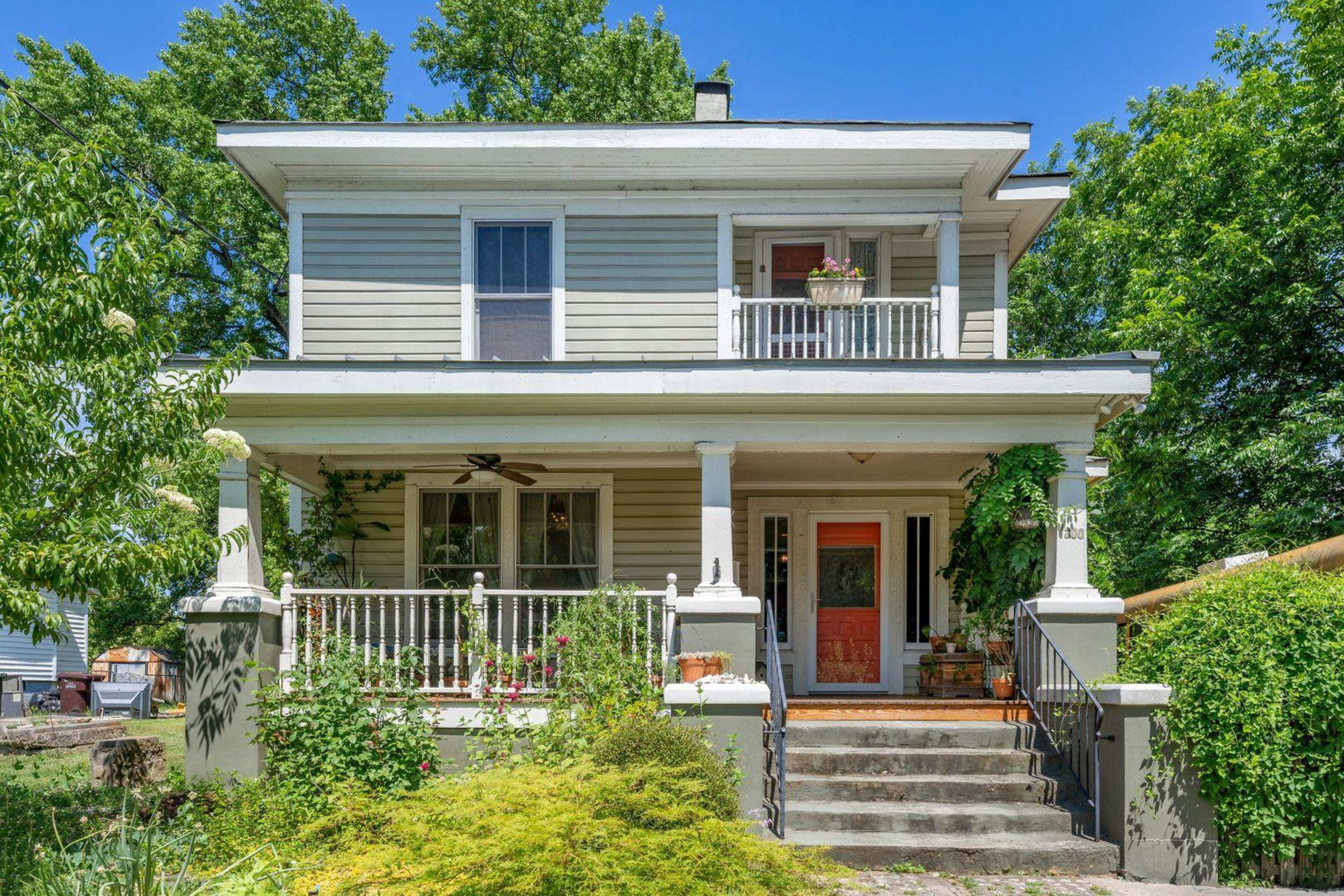 The image shows a two-story house with gray siding, a front porch supported by white columns, and lush greenery surrounding the entrance.