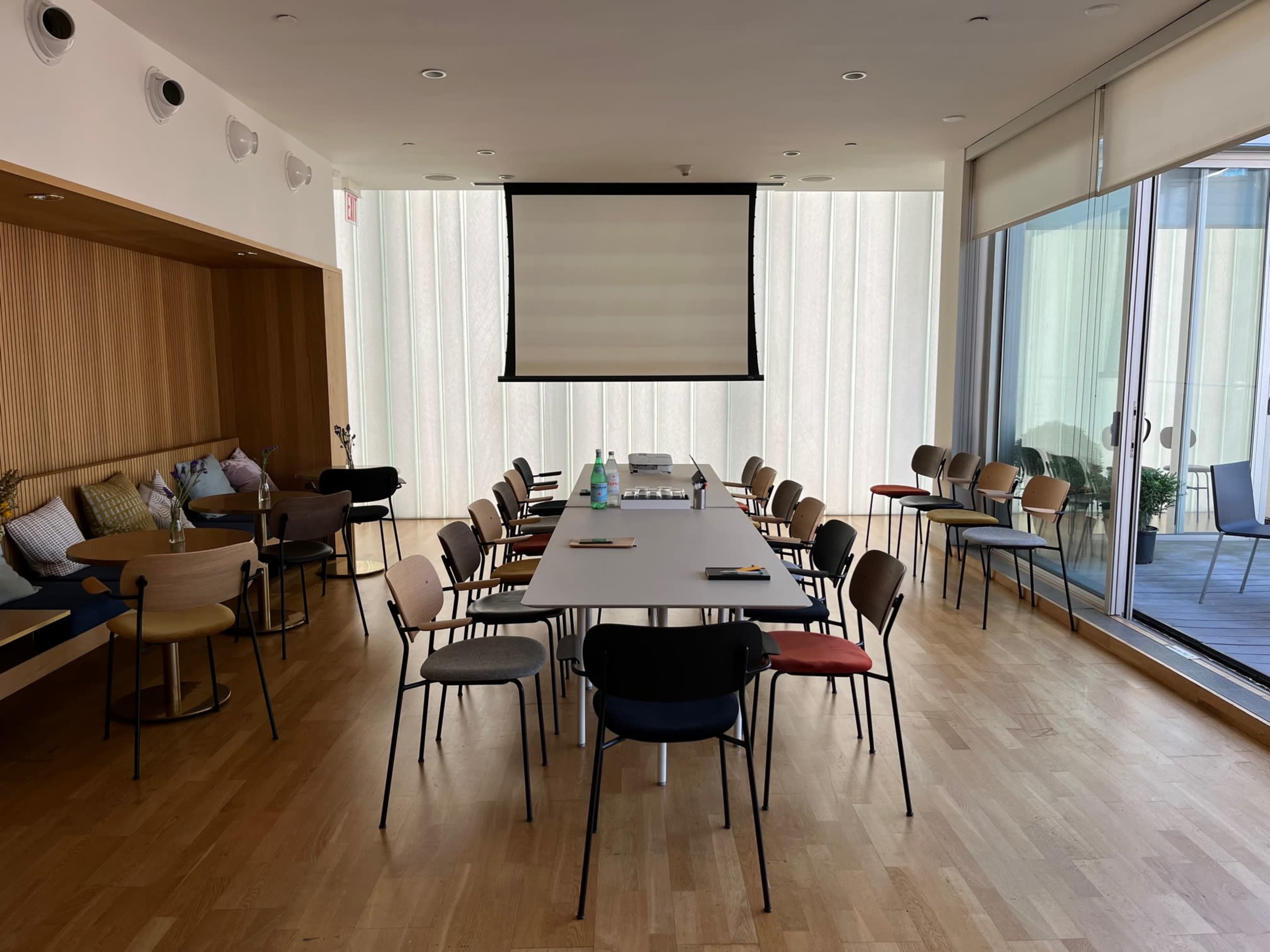 A conference room is set up with a long table surrounded by chairs, facing a screen, with natural light streaming through large windows.