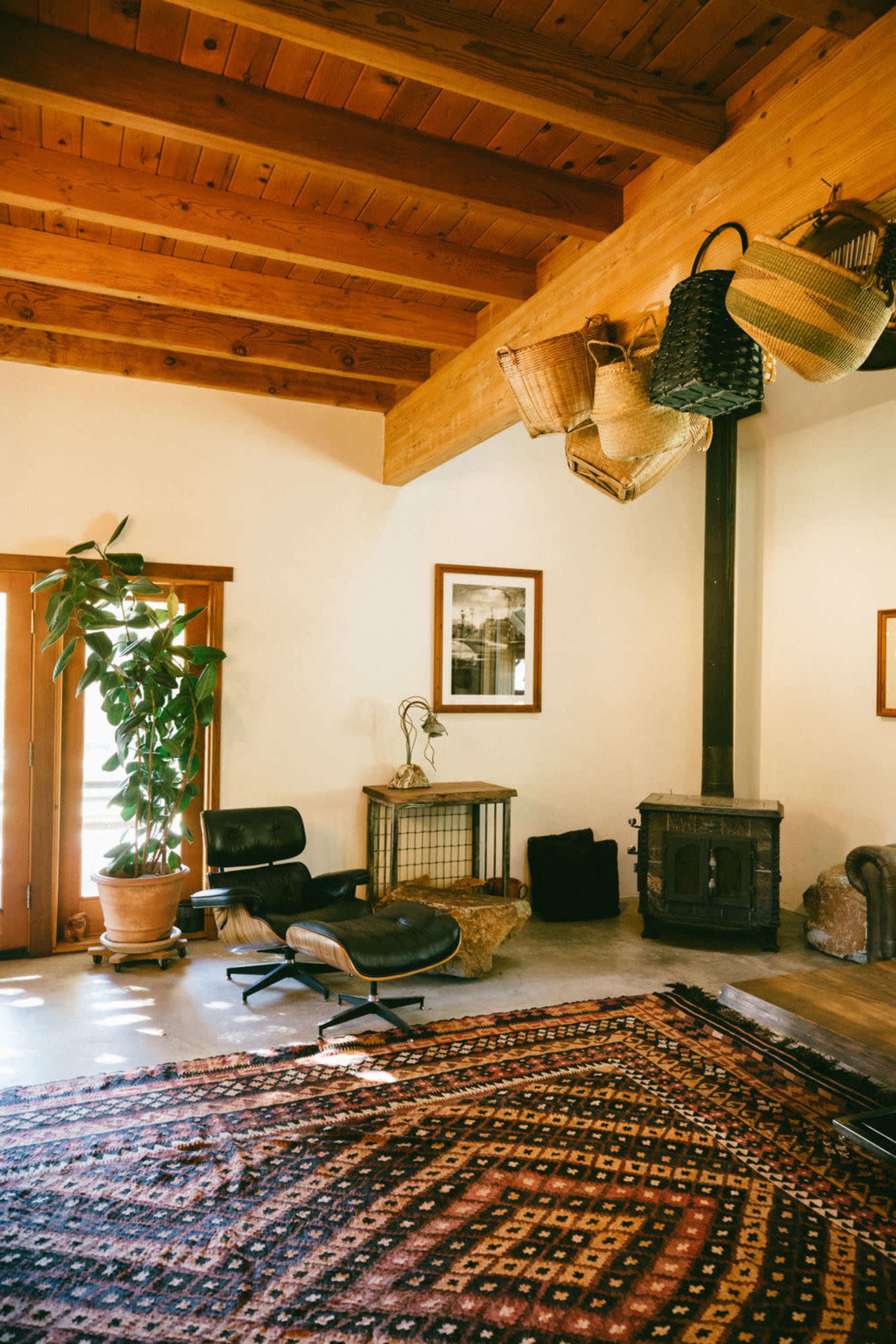 A cozy living room features wooden beams, a large area rug, potted plants, and several hanging baskets.