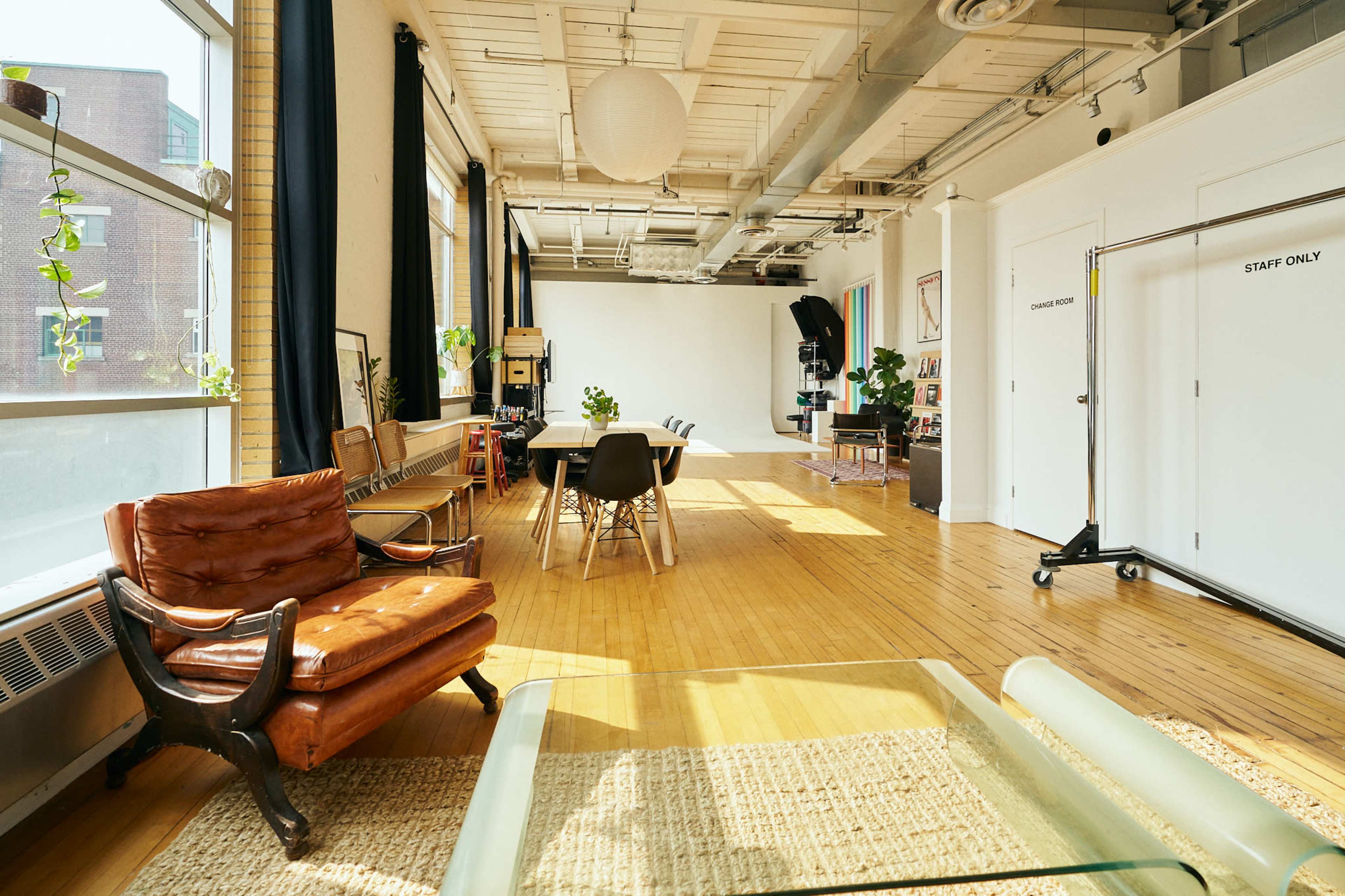 A spacious, well-lit studio features wooden floors, a long table surrounded by chairs, and a bright backdrop area for photography, with plants and minimalist decor.