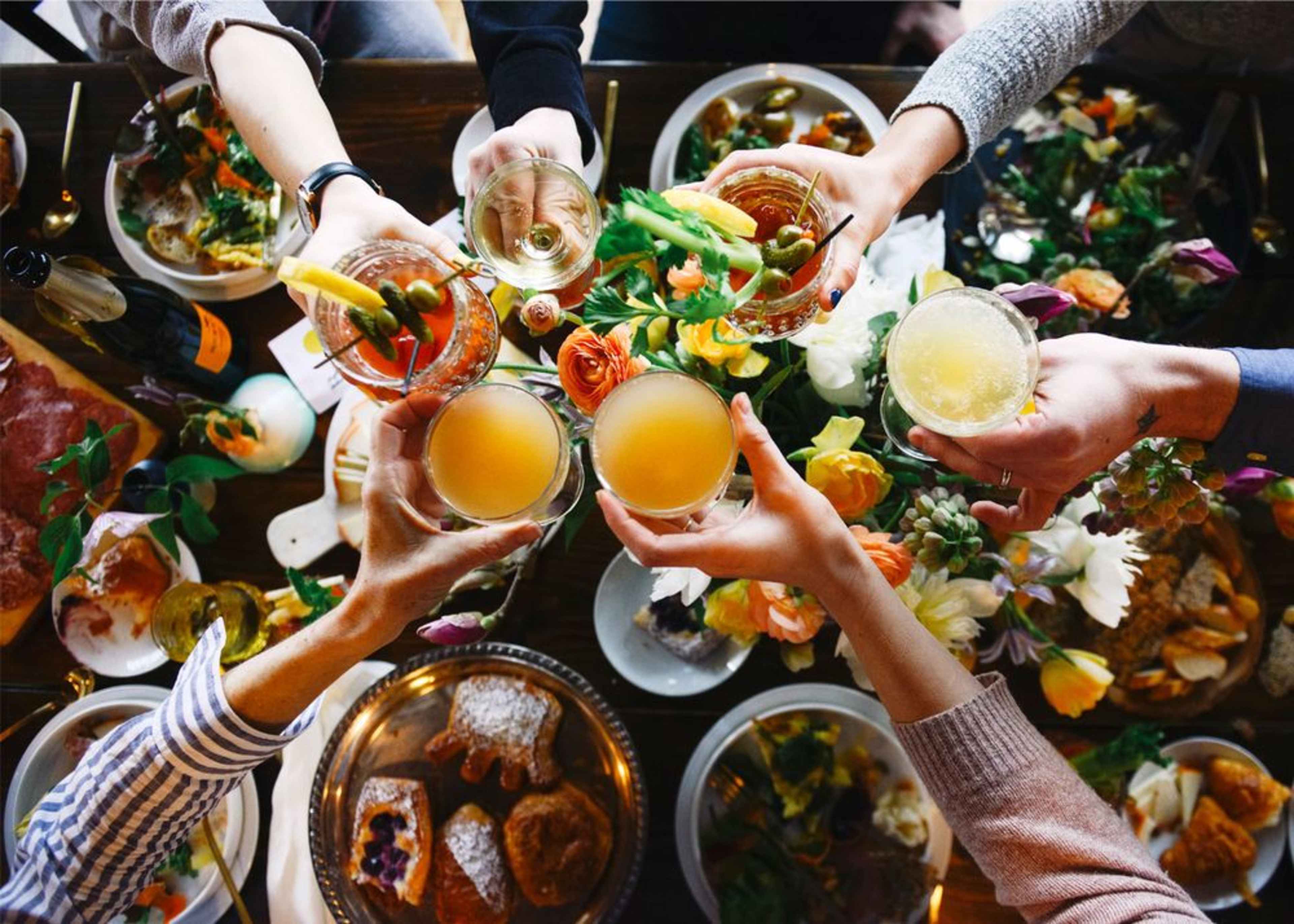 A group of hands raises various drinks in a toast above a table set with food and floral decorations.