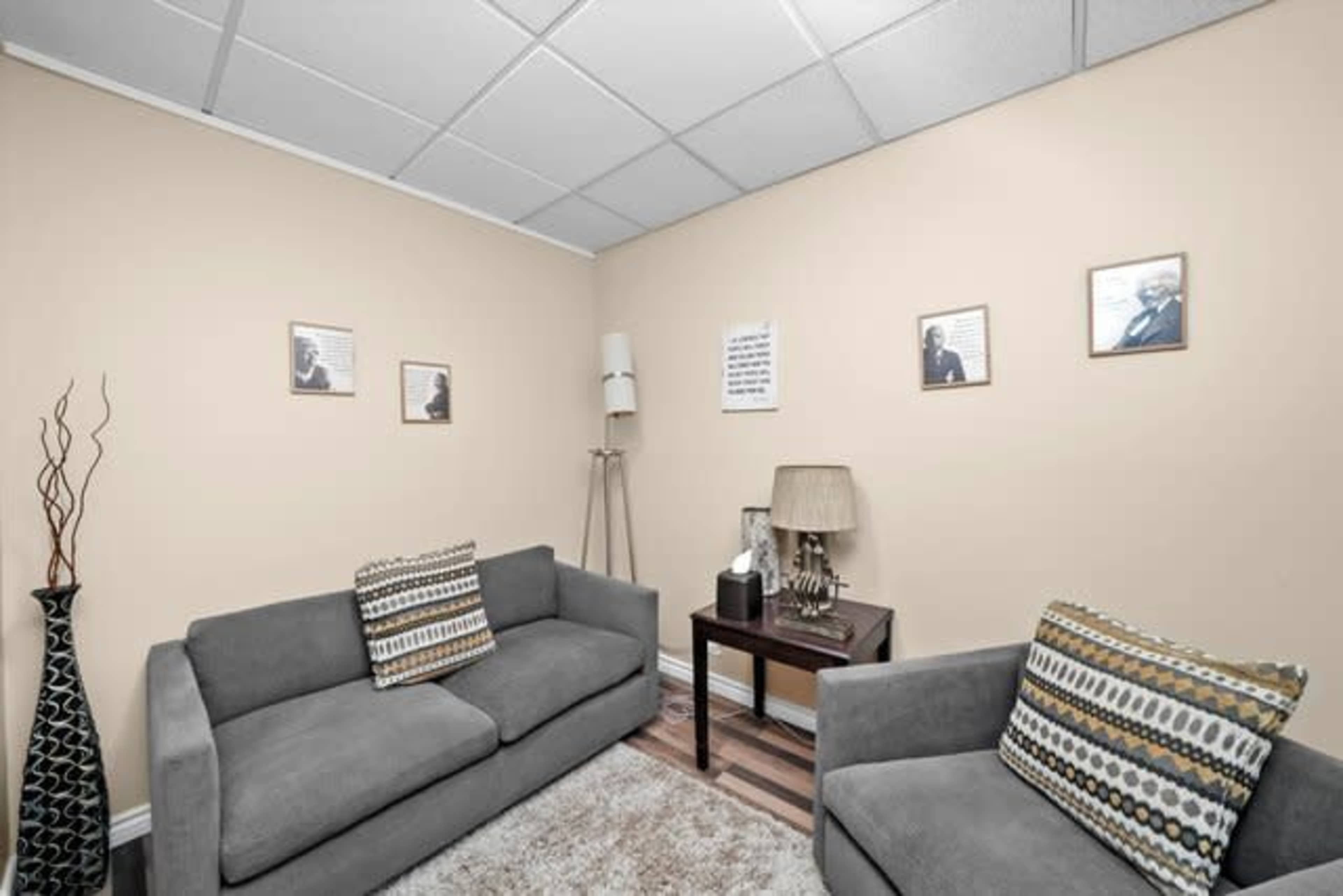 The image shows a small, neatly arranged room with two gray sofas, a side table with a lamp, and framed photographs on the walls.