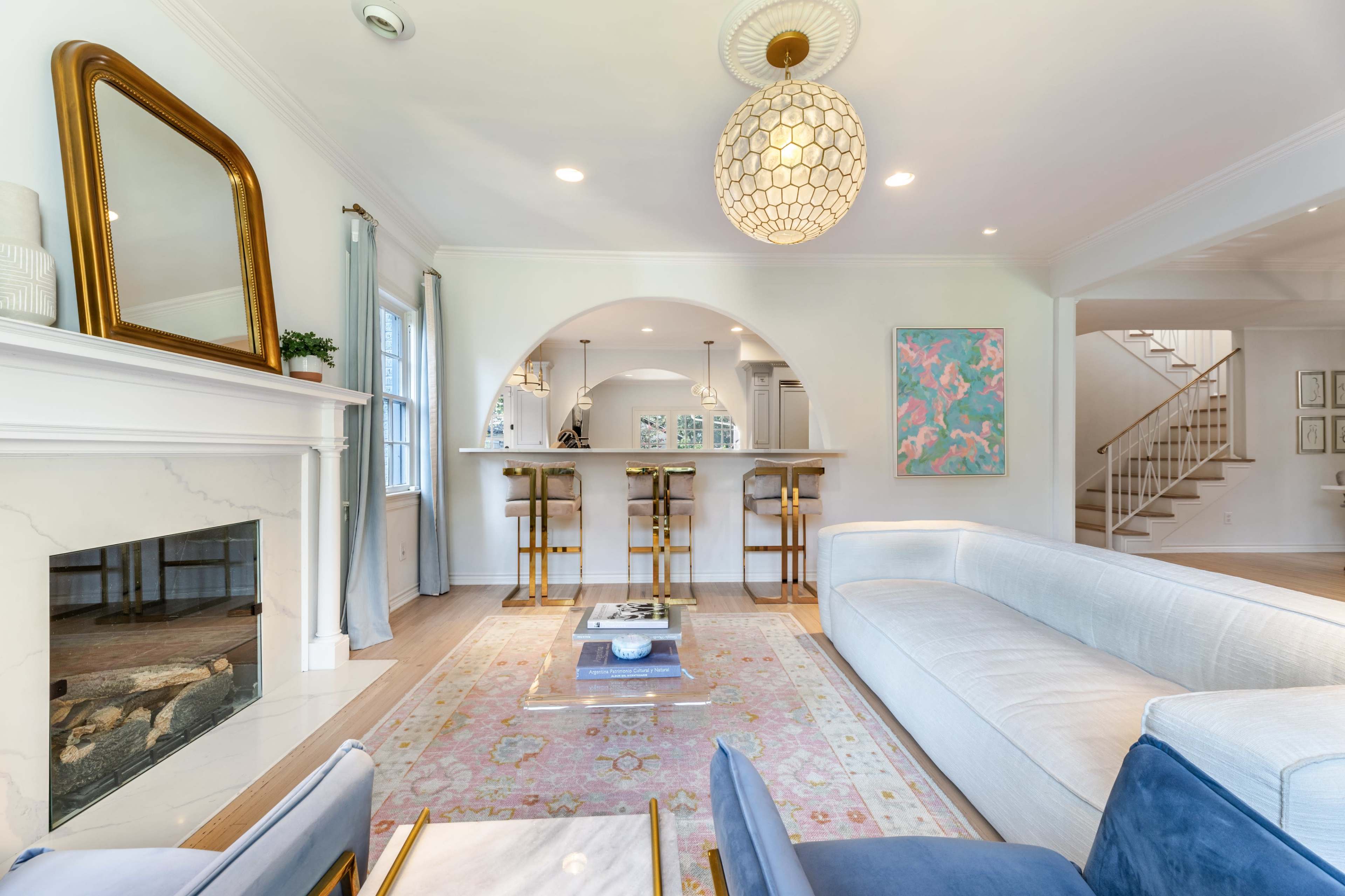 The image shows a bright living room with a fireplace, a blue sofa, a coffee table on a patterned rug, and a bar with high stools in the background.