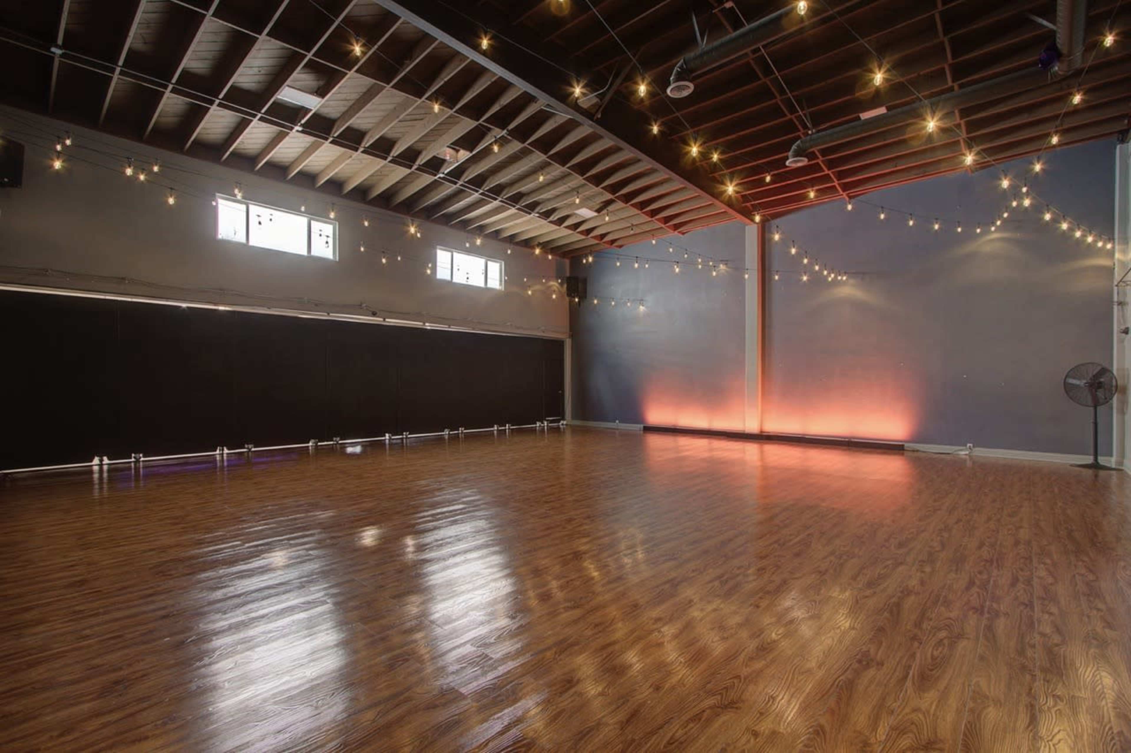 A spacious dance studio features polished wood flooring and soft lighting, with large windows and a black wall at one end.