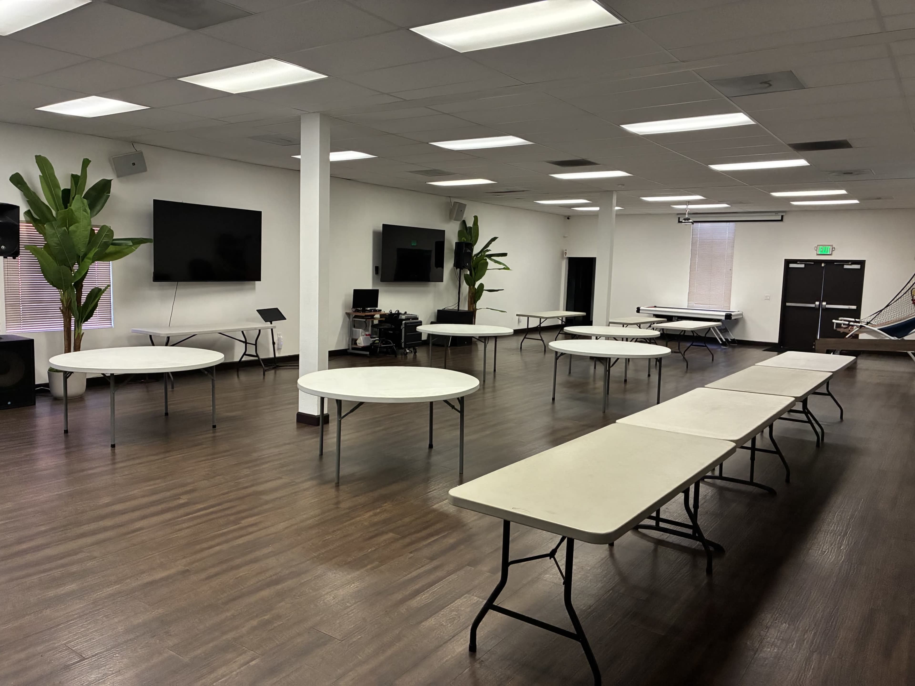 The image shows a spacious room with several round and rectangular tables arranged throughout, along with potted plants and two wall-mounted screens.