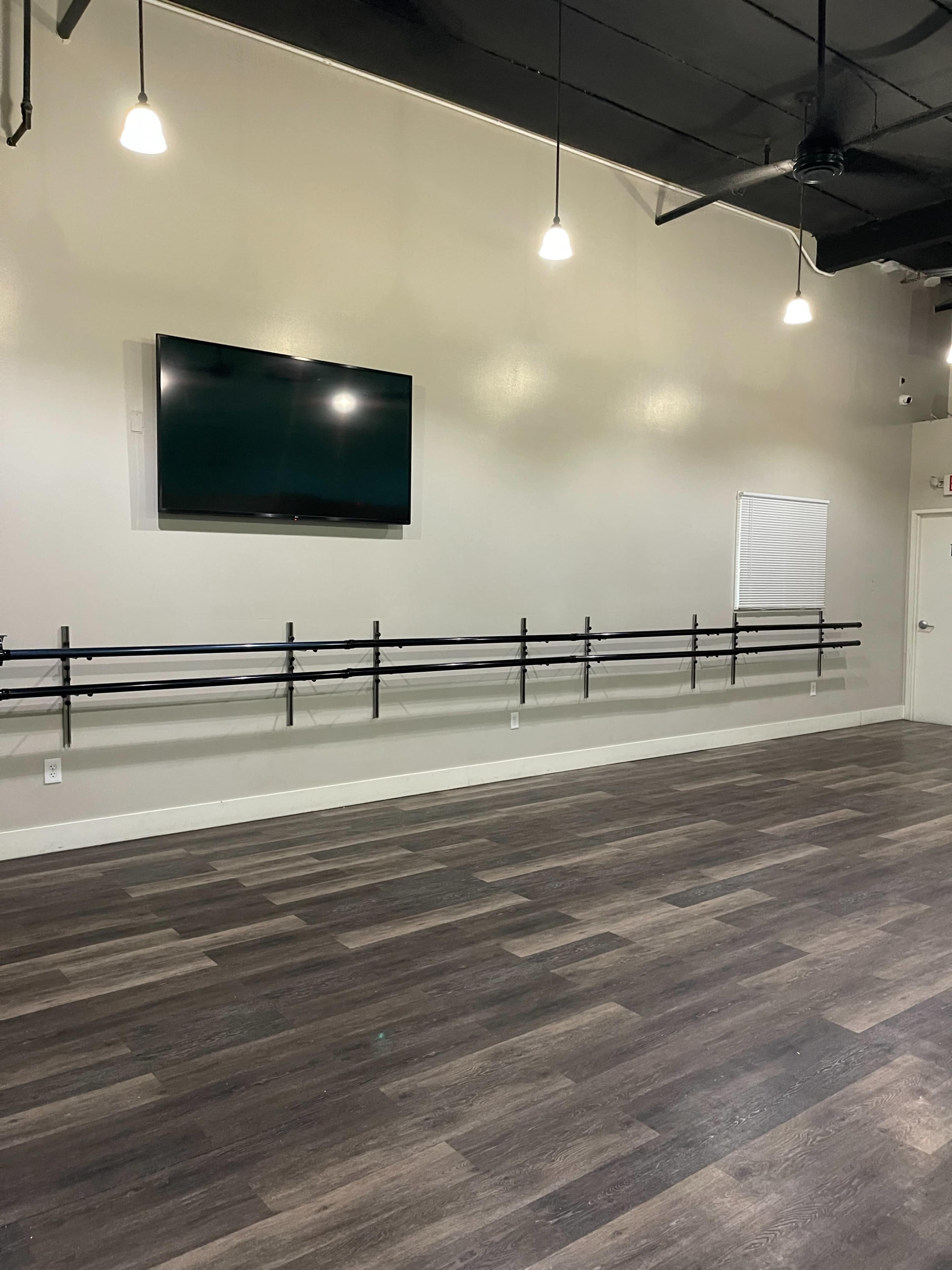 An empty dance studio featuring a wall-mounted television and a horizontal ballet barre along one wall.