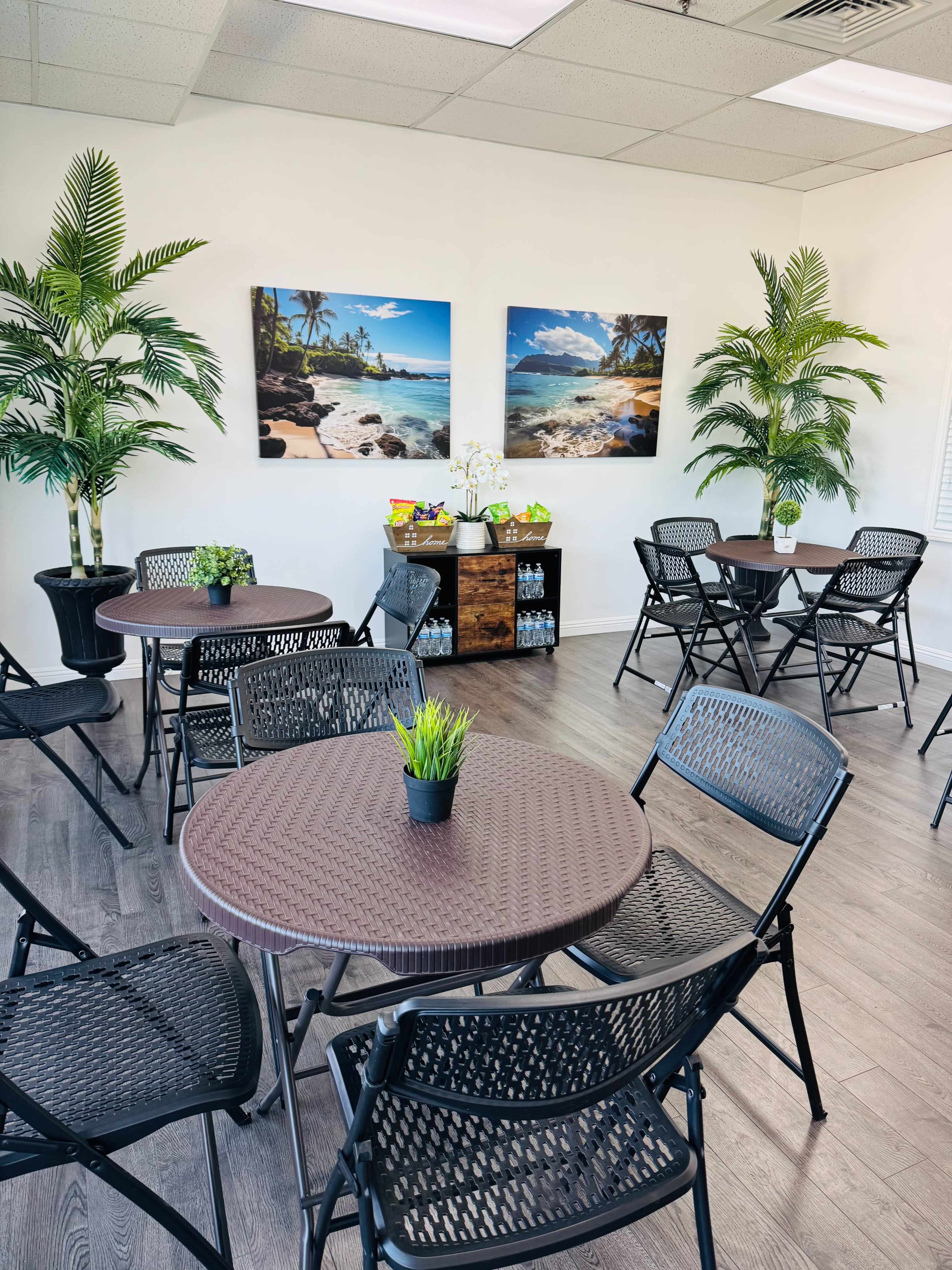 The image shows a bright, organized room with several round tables and chairs, adorned with potted plants and tropical-themed wall art.