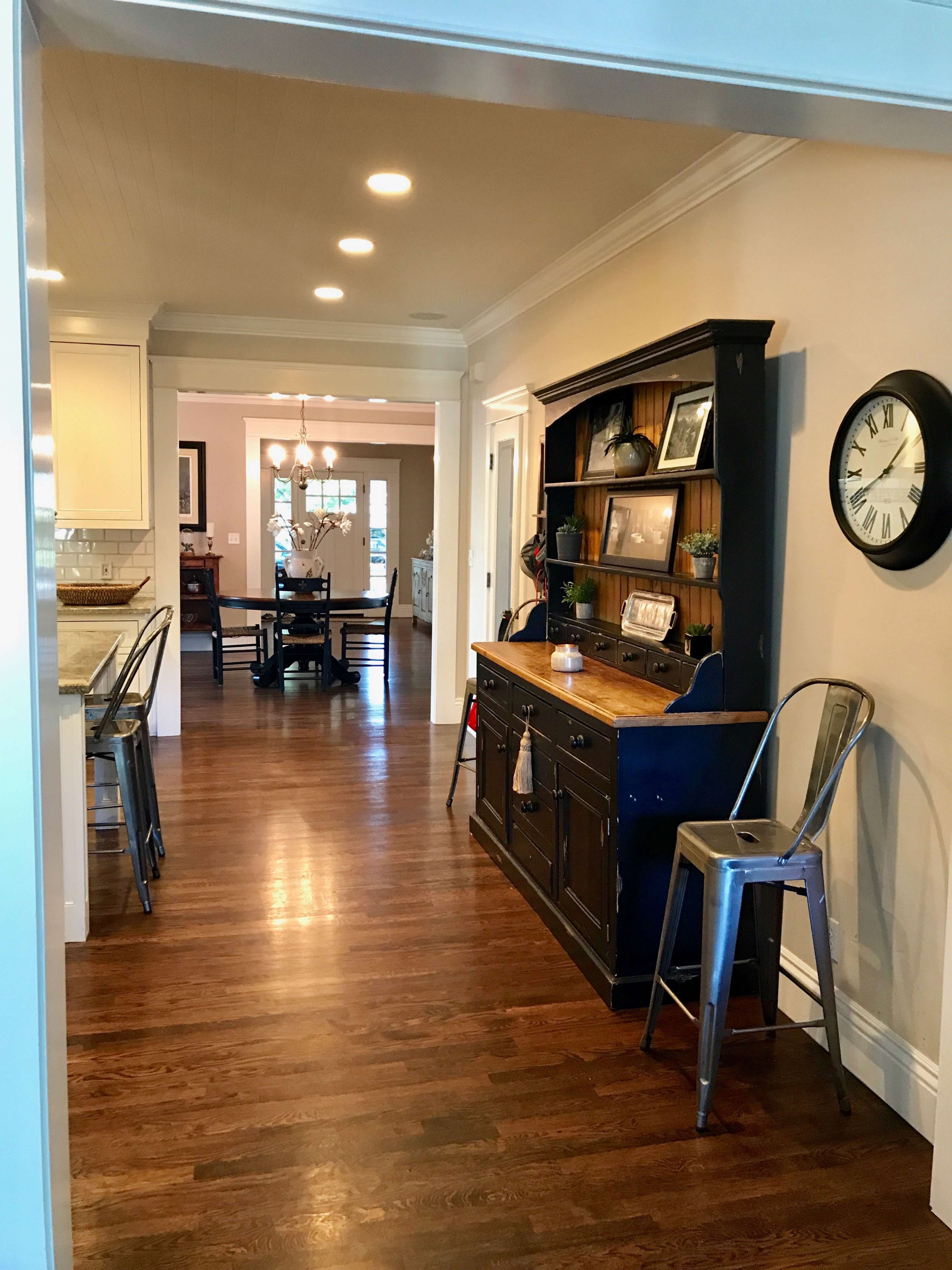A hallway connects a kitchen to a dining area, featuring a dark wood sideboard and a clock on the wall.