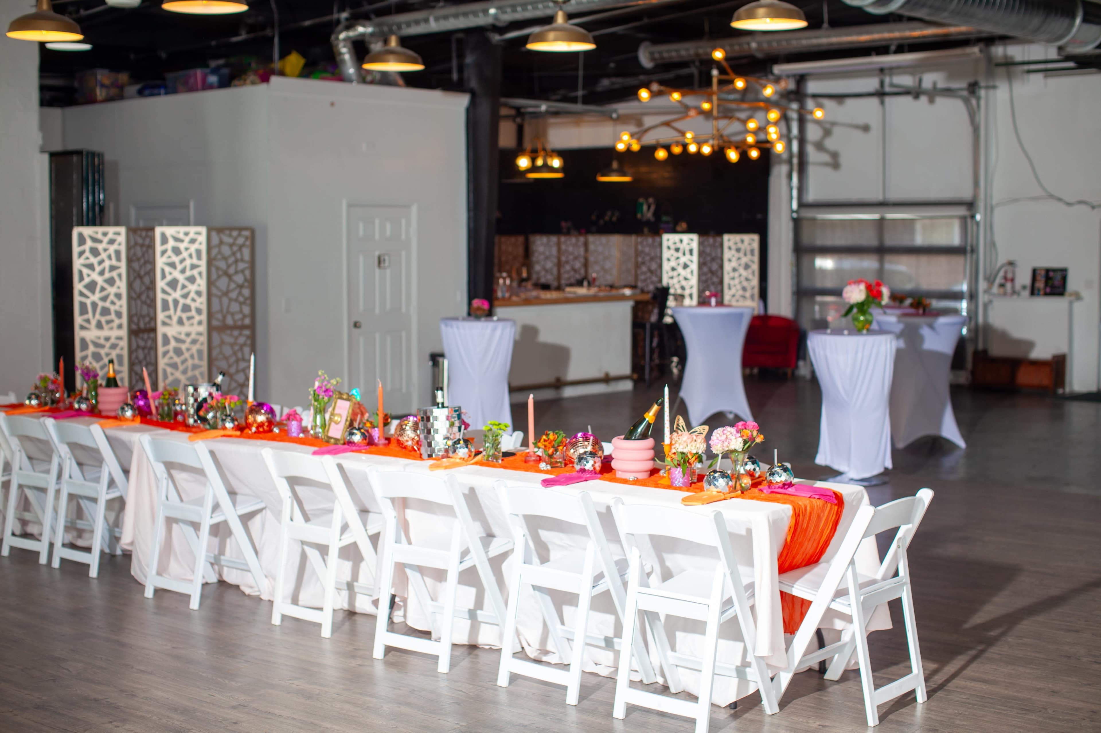 A long rectangular table is set with colorful decorations and flowers, surrounded by white chairs in a well-lit event space.