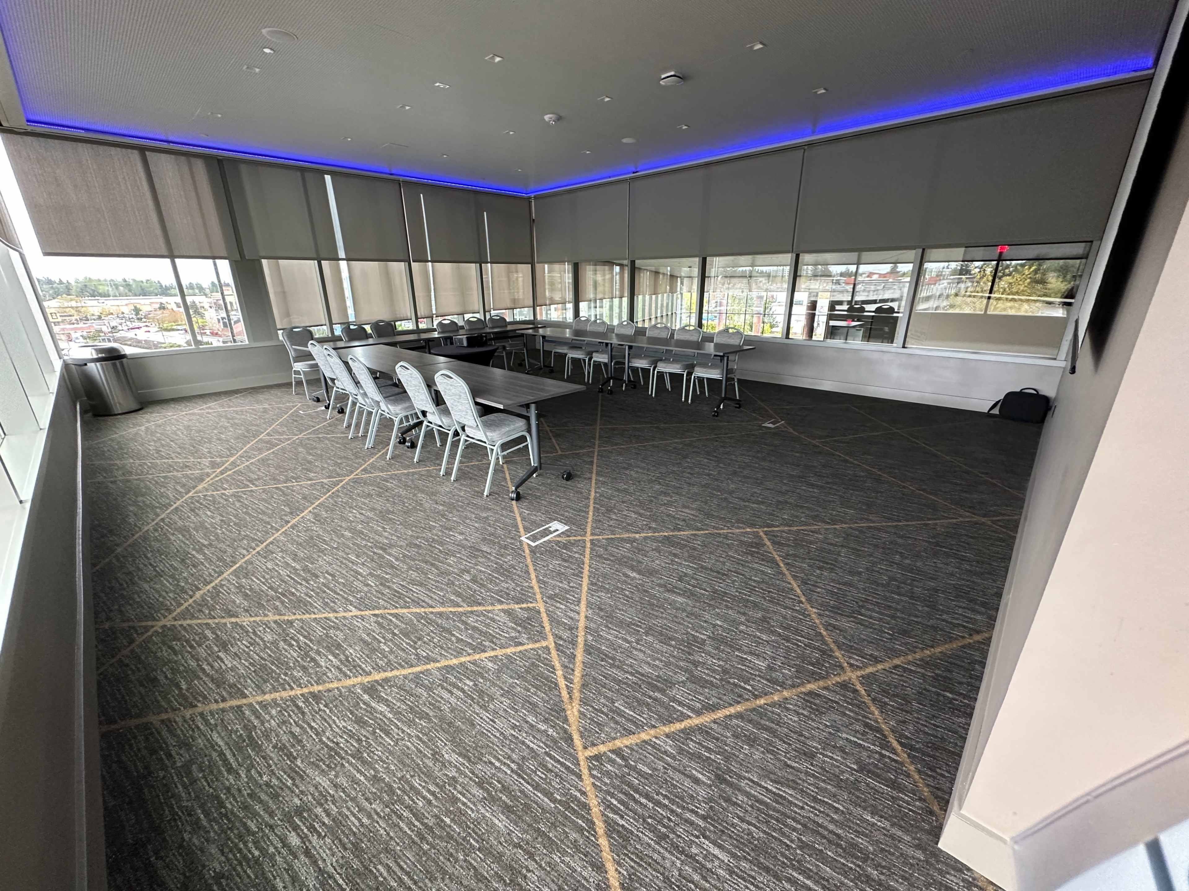 The image shows a modern meeting room with several rows of chairs around a large rectangular table and large windows along one wall.